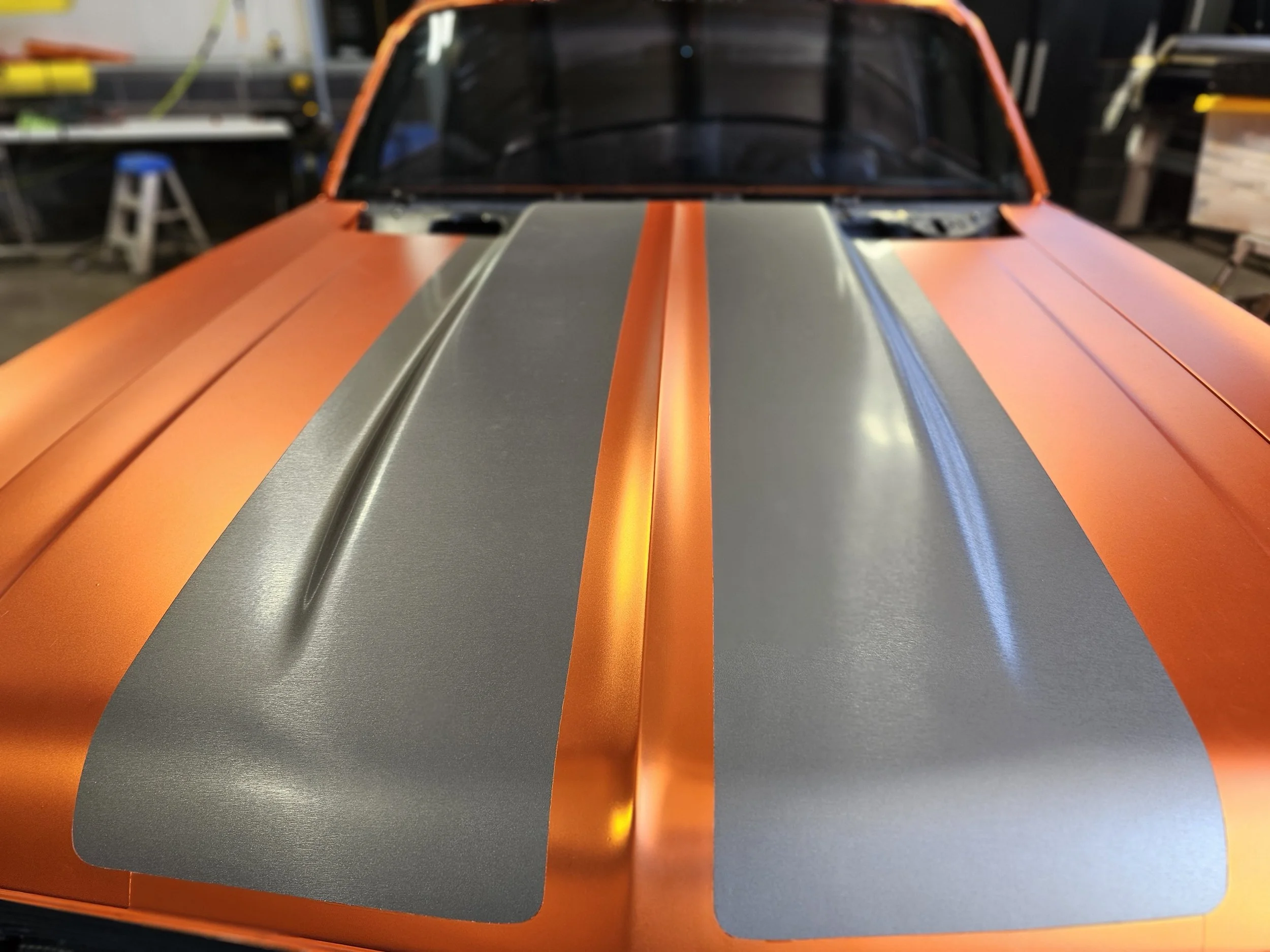 Close-up view of a race car with orange and silver paint, showing the hood with racing stripes in black, orange, and yellow, in a garage or workshop setting.