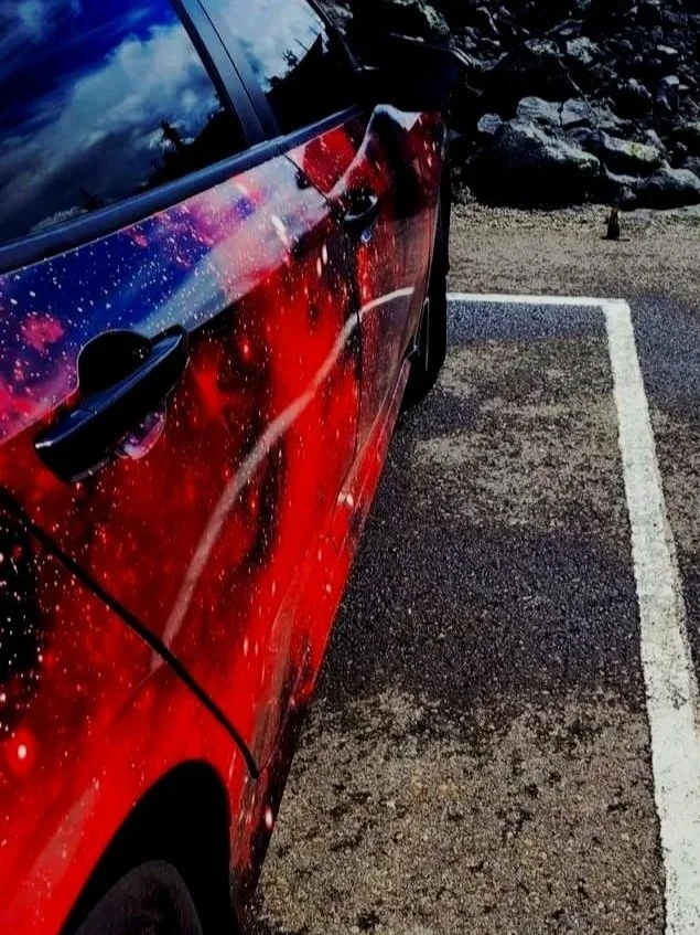 Red car with galaxy-themed vinyl wrap parked in a marked parking spot on asphalt, with rocks and gravel nearby.