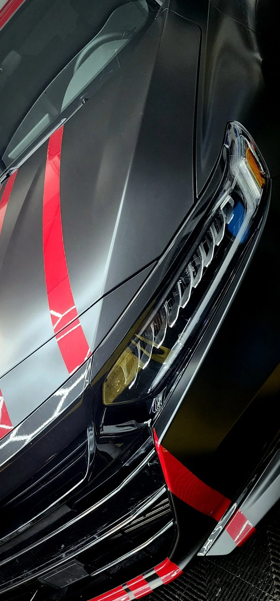 Close-up of a black sports car with red racing stripes, showing the front headlight, part of the hood with a red stripe, and the windshield.