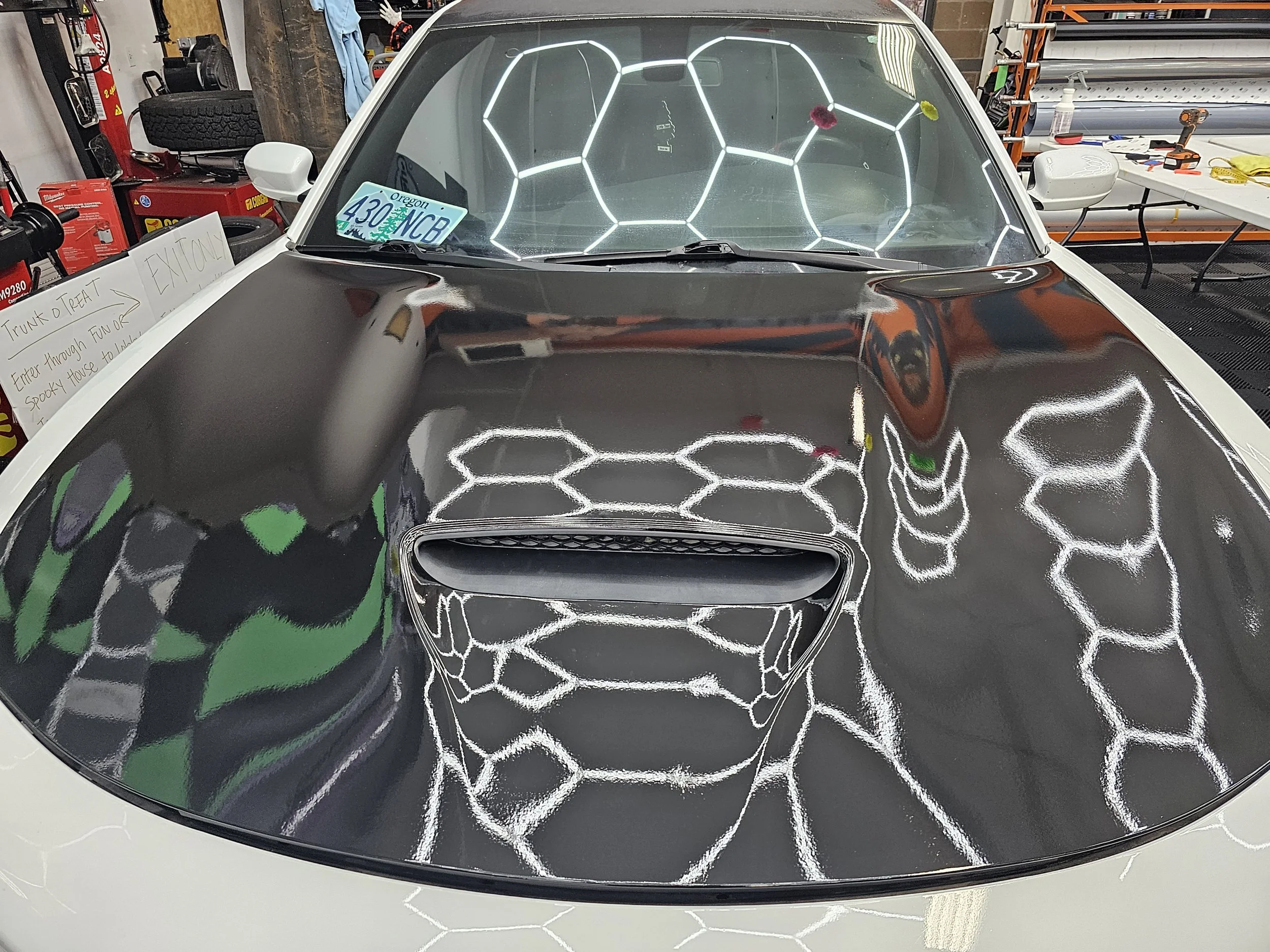 The hood of a white sports car with a black section featuring a custom honeycomb and skull graphic design. The windshield displays neon-like geometric accents, with a small Oregon license plate on the passenger side. The garage background includes tools, shelves, and a sign.