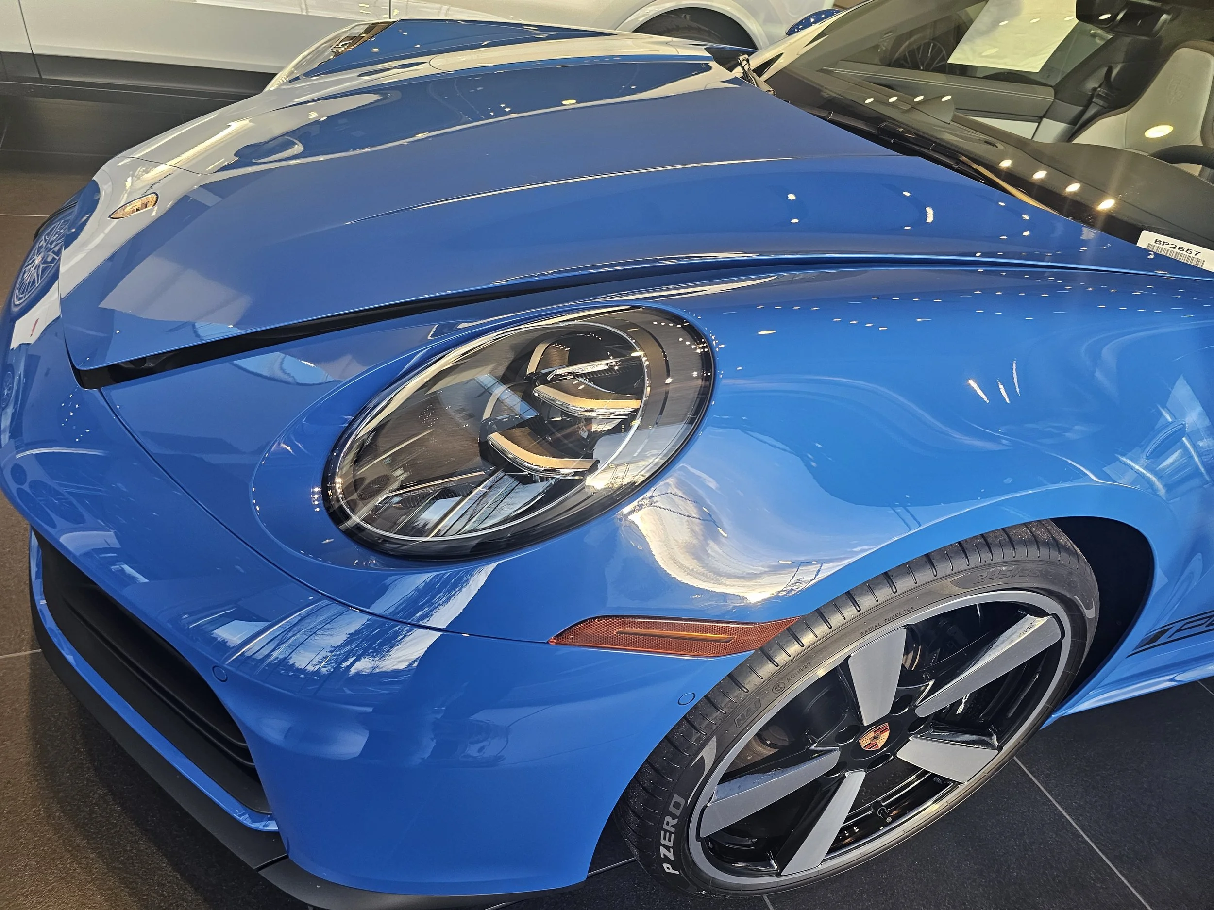 Close-up of a bright blue Porsche sports car showing its front and side, including the headlight, tire, and part of the windshield, parked indoors.