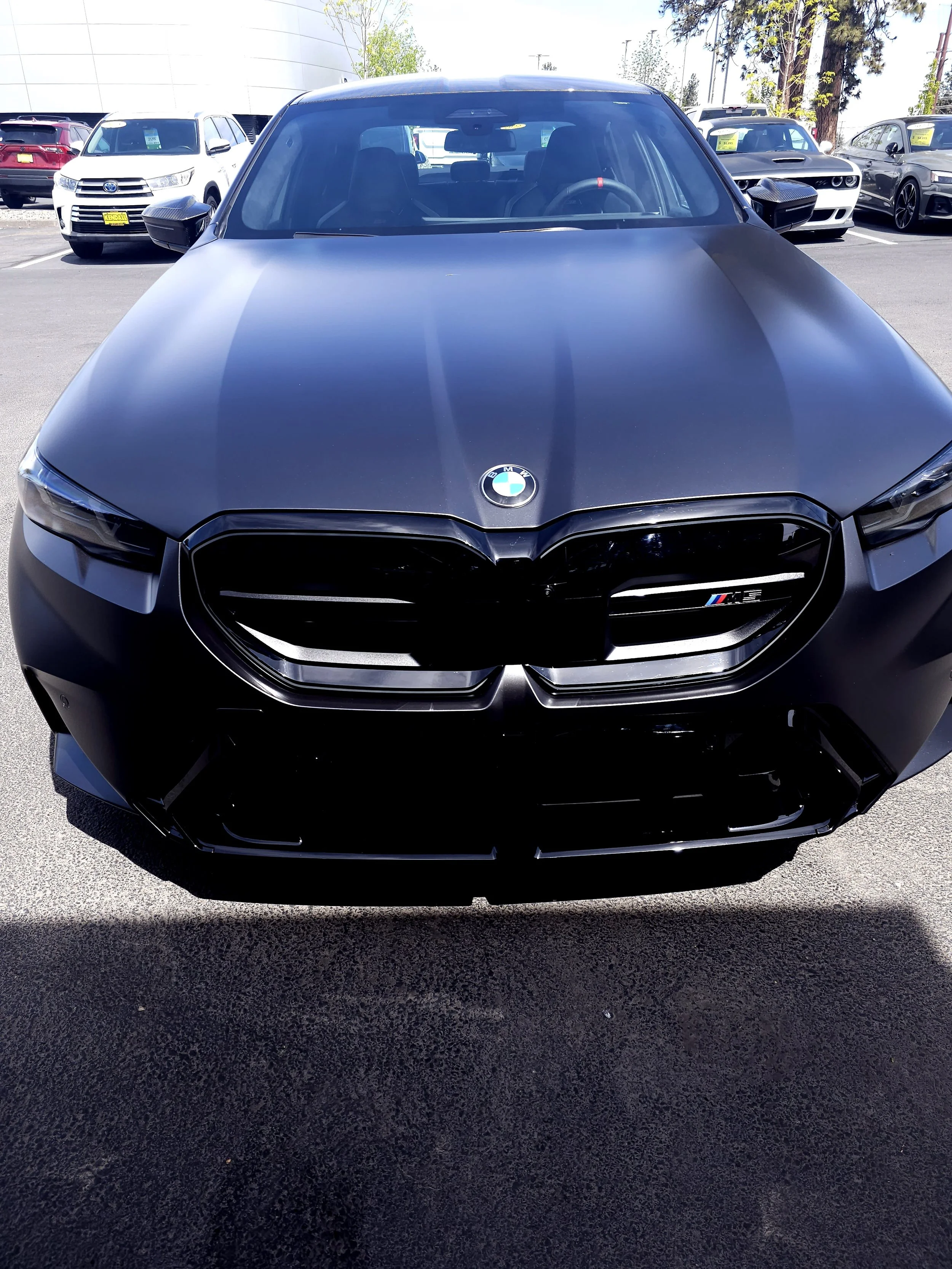 Front view of a black BMW car with an M badge on the grille in a parking lot, with other cars and trees in the background.