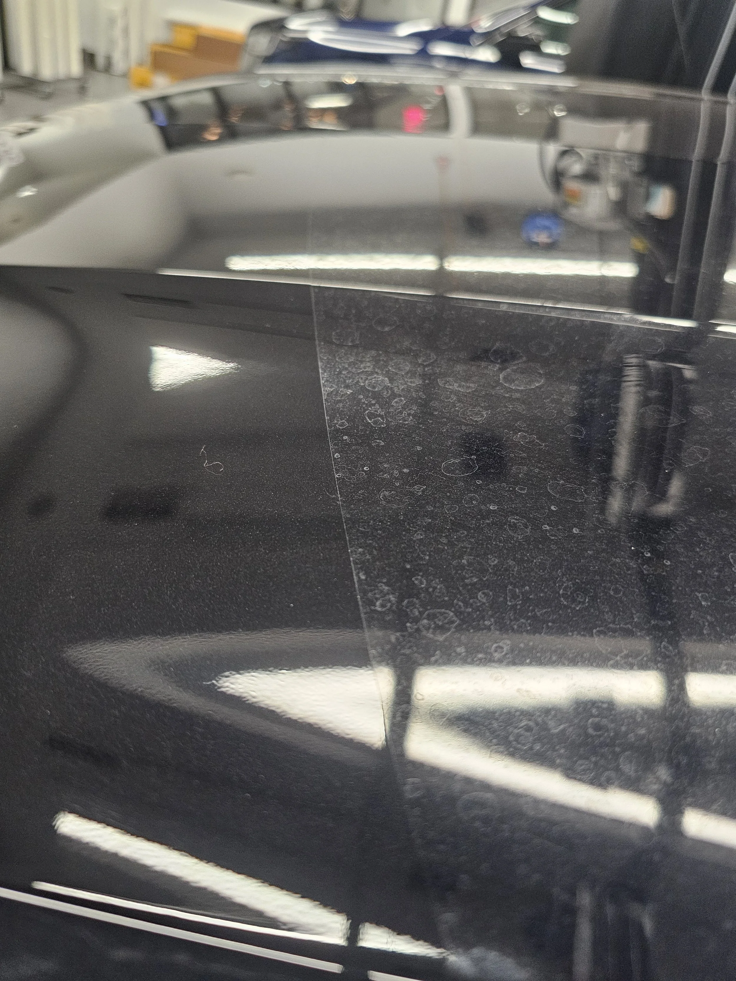 Close-up of a black car with a shiny, reflective surface, parked indoors, with ceiling lights and other cars visible in the reflections.