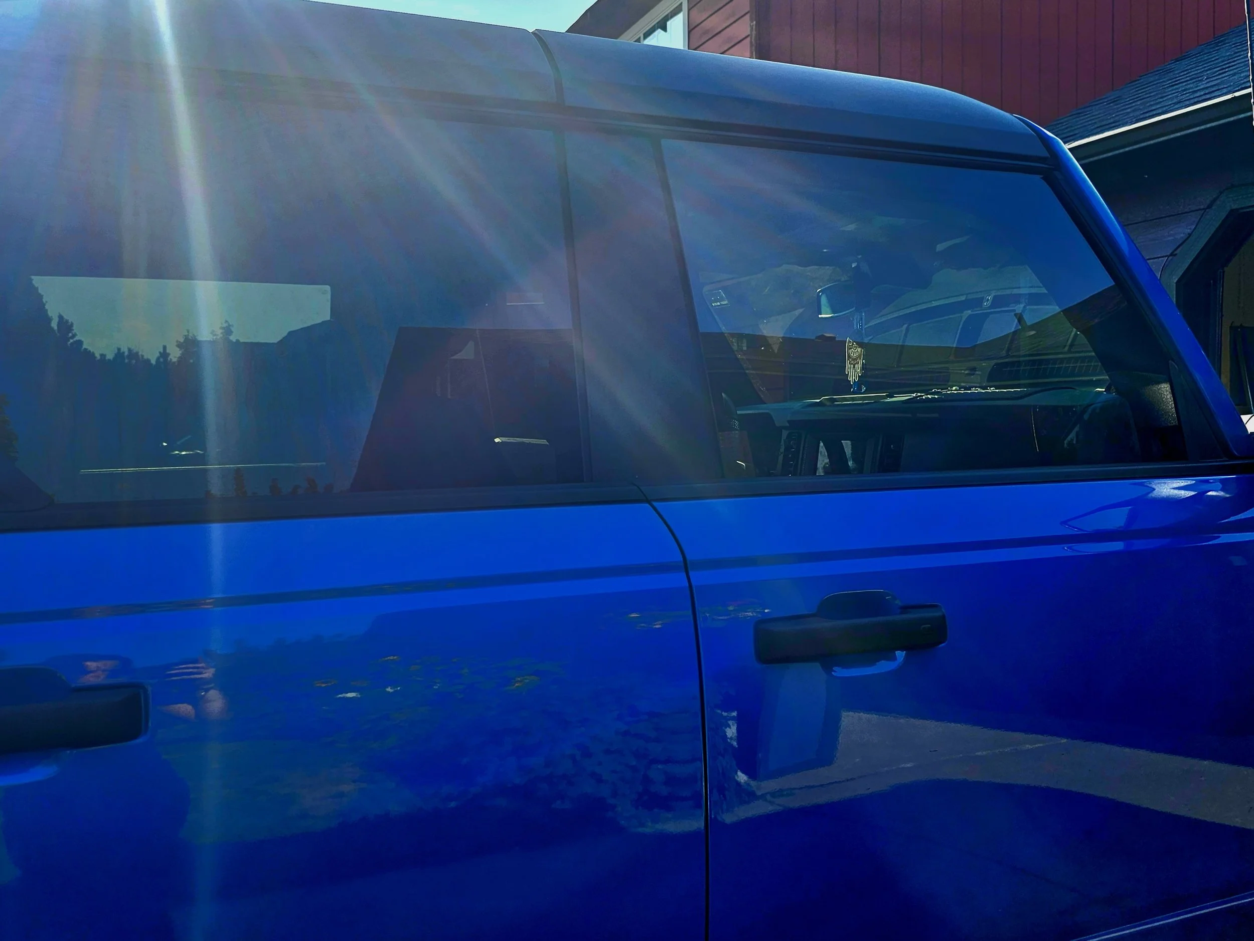 Close-up of the front passenger side of a blue van parked outside a house with a red exterior. The van's windshield and part of the dashboard are visible, with reflections of the sky and surroundings on the glass.