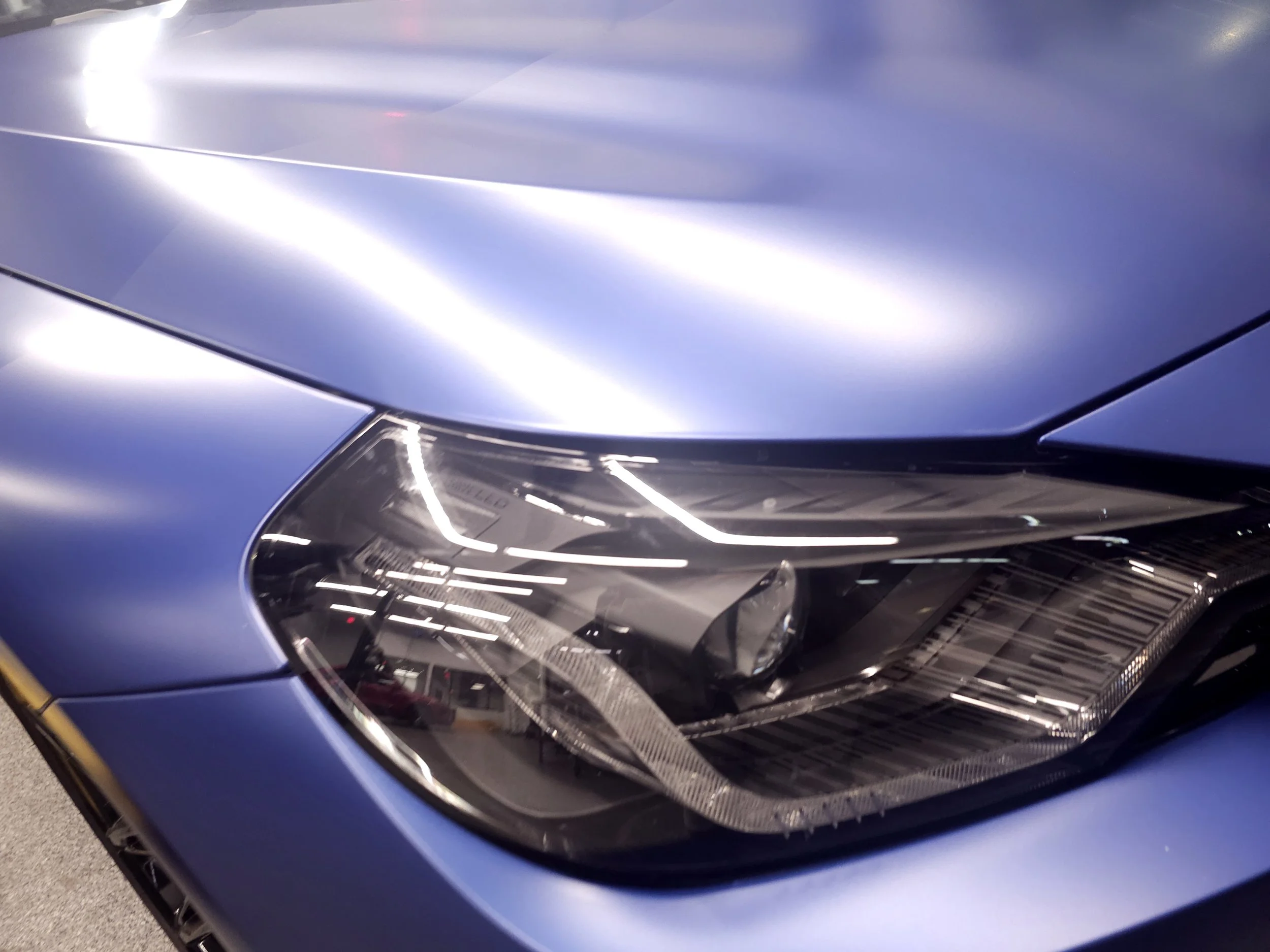 Close-up of a blue car's headlight and front end, showcasing its sleek design and reflective surface.