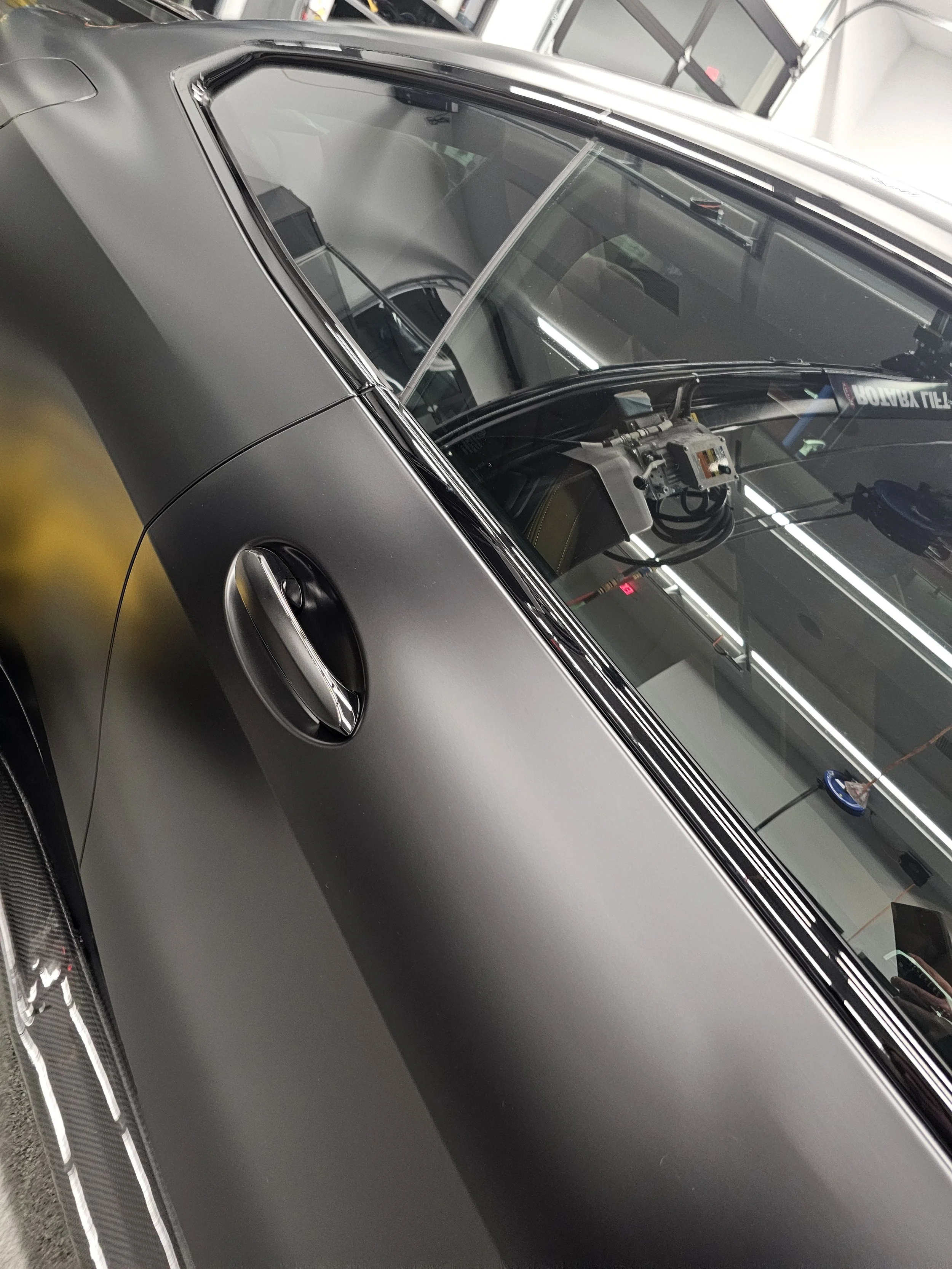 Close-up view of a modern black sports car's windshield, roof, and part of the hood with a sleek design and air intake vent, parked indoors.