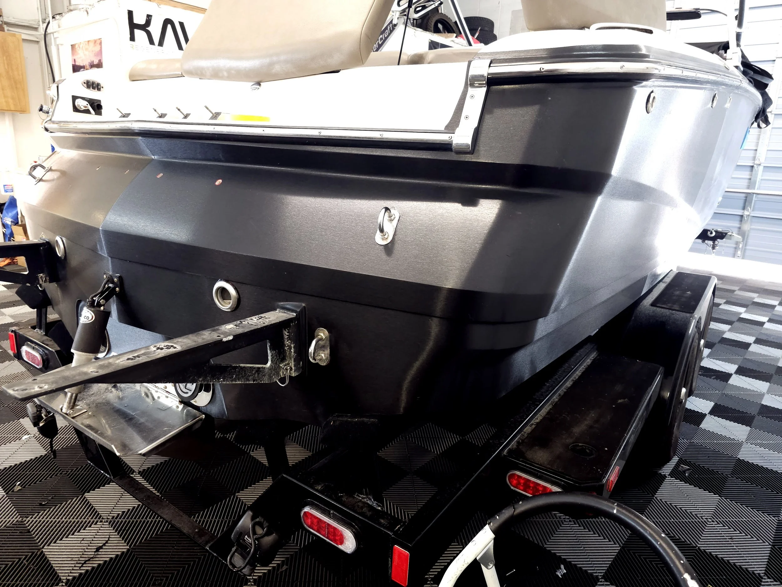 Black boat on a trailer inside a garage. The boat features a beige seat and metal fixtures along the side, with a patterned black and gray floor.