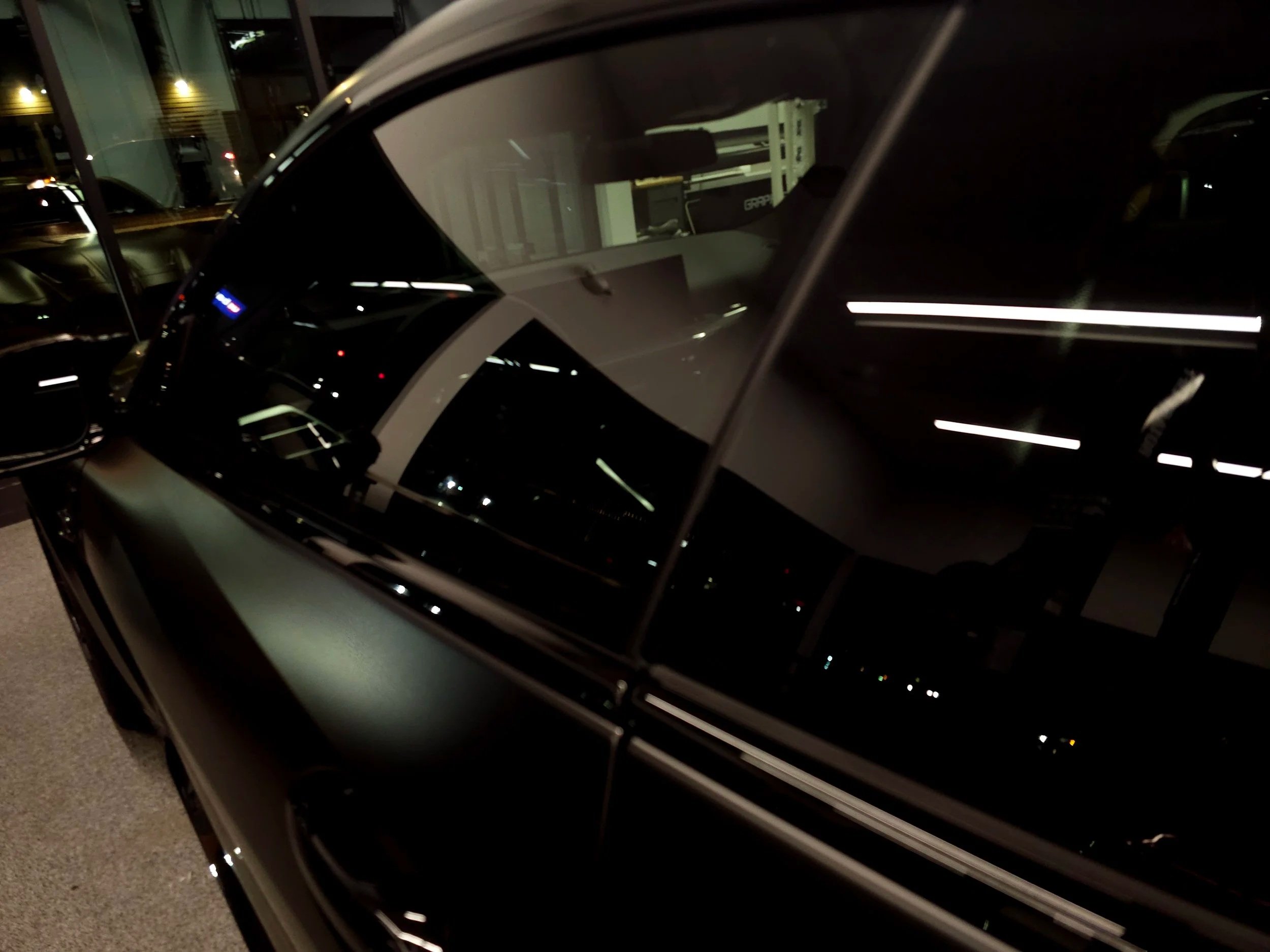 A black car parked indoors with reflections of overhead lights on its surface, and a building exterior visible through the windows.