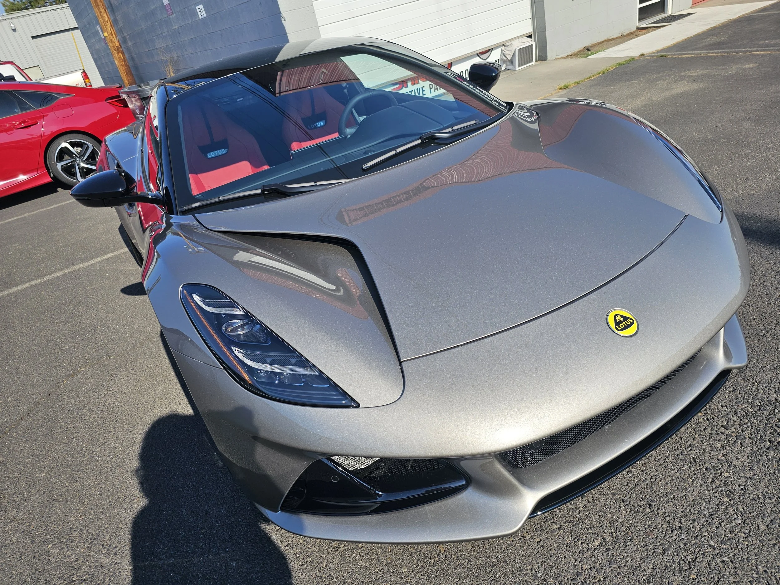 Gray sports car with a Lotus logo on the hood, parked outdoors next to a red car, in a parking lot with a gray building behind.
