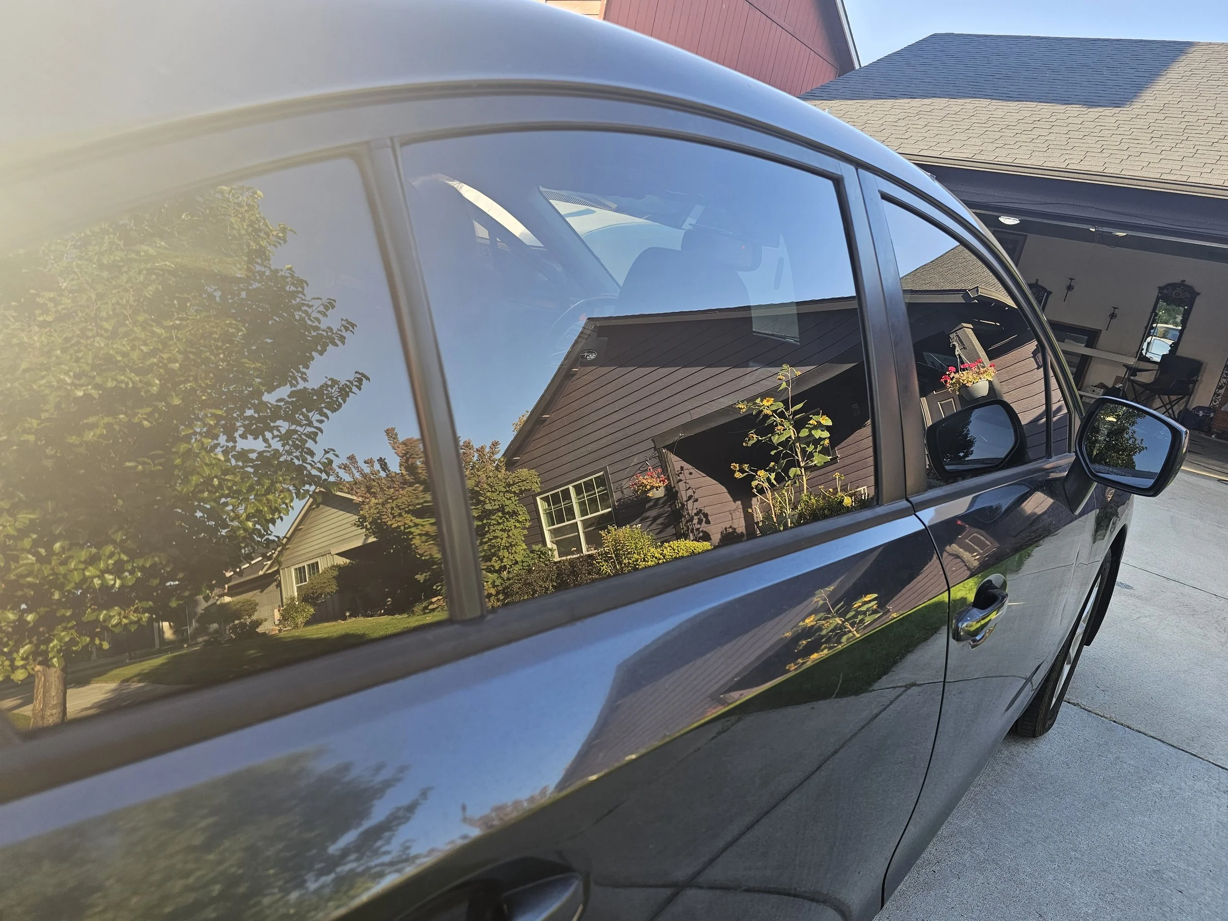 Reflection of a house, trees, and sky on the shiny black side of a car parked in a driveway.