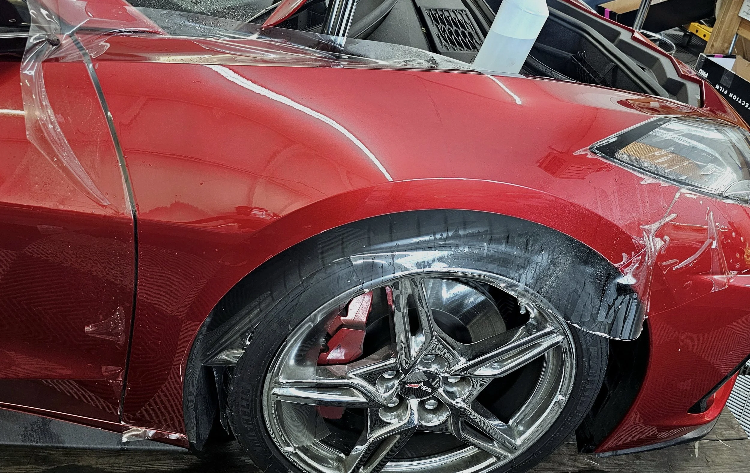 Close-up image of a red sports car undergoing repair or detailing. The front wheel is visible with a shiny chrome rim, and the car's body has protective plastic film on the side panel. The background shows workbenches and tools typical of an auto repair shop or detailing garage.