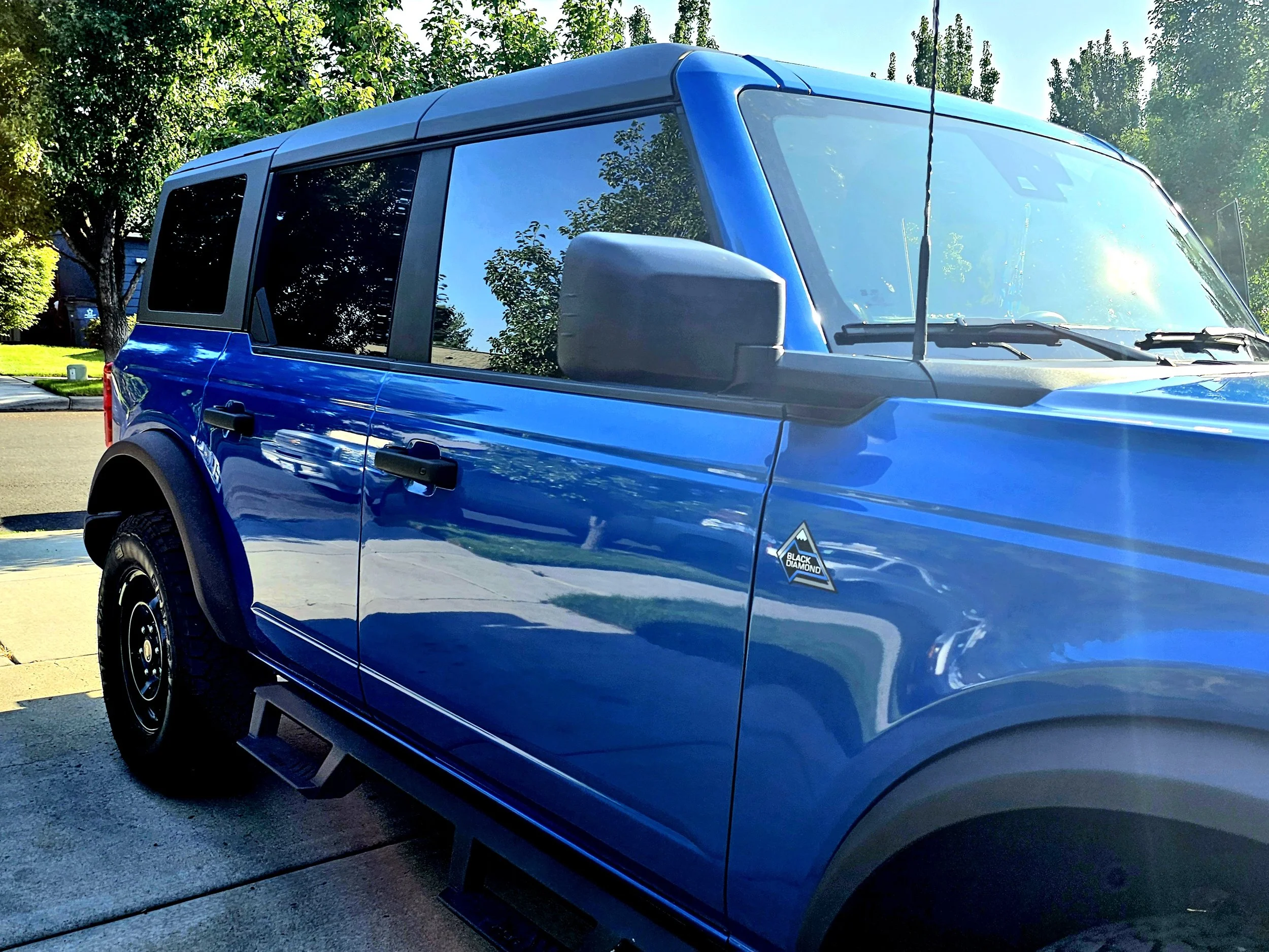 Close-up of a blue SUV parked on a suburban street, with trees and houses in the background, and sunlight reflecting off the vehicle.