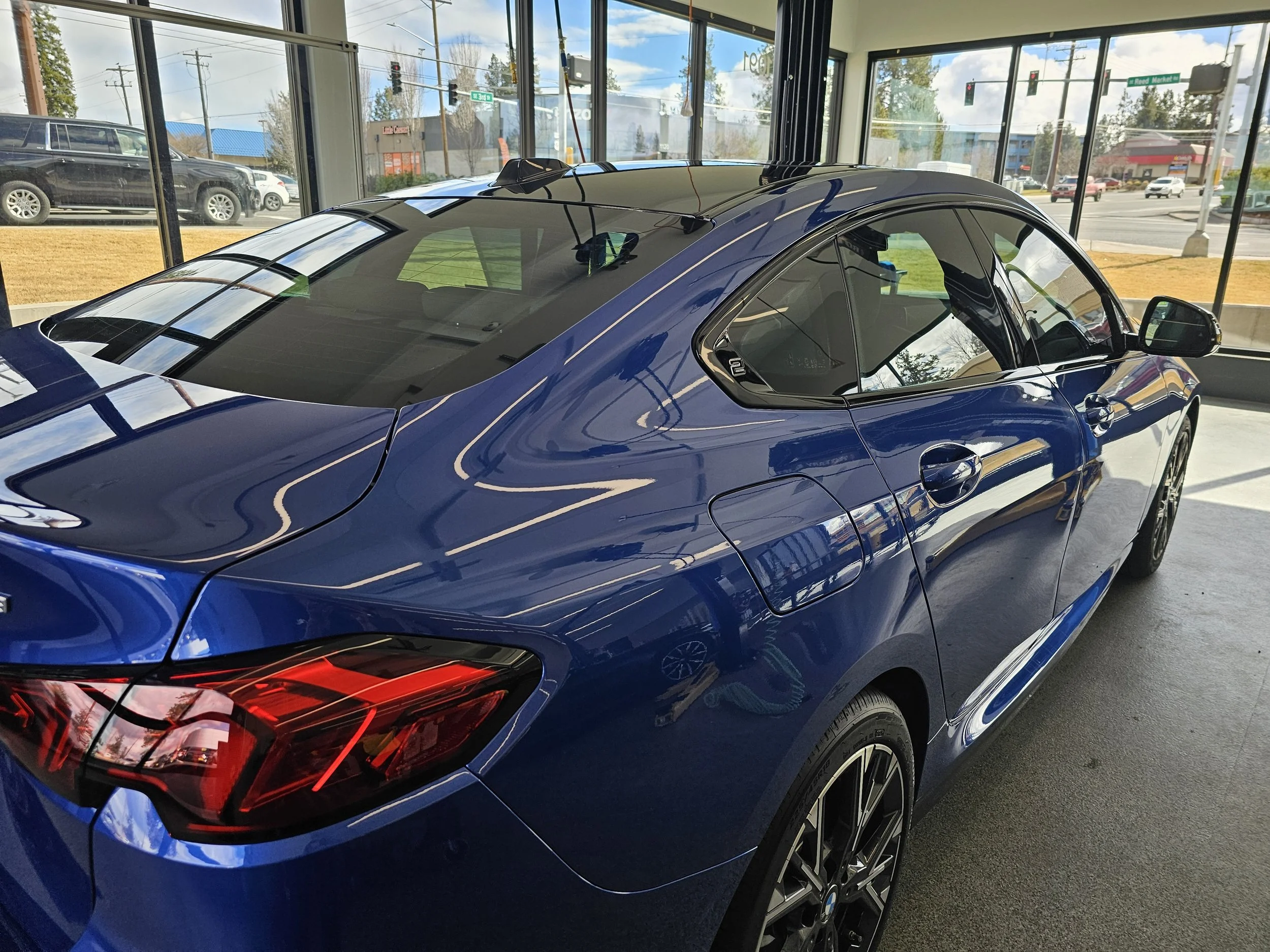 A blue sports car with sleek lines and red taillights is parked inside a showroom with large glass windows, reflections on the car's surface, and a view of the street outside.