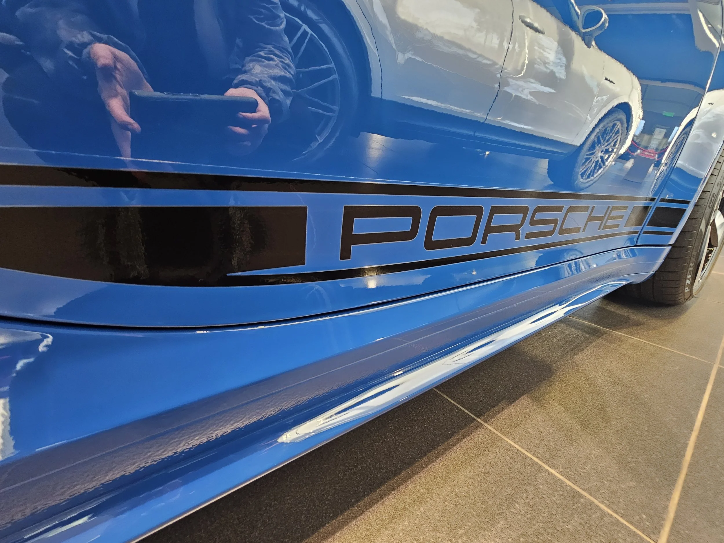 A blue Porsche car with the Porsche logo visible, parked indoors on a tiled floor. A person is reflected on the car's surface, holding a phone to take the photo.
