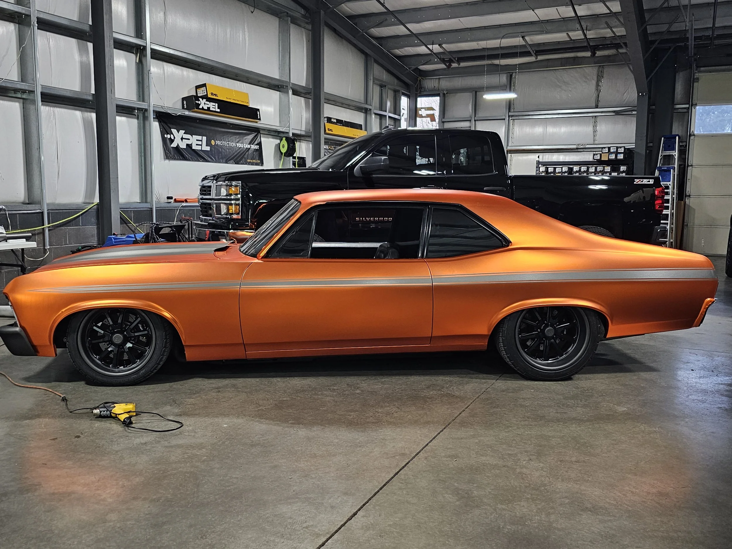 An orange classic muscle car with black wheels parked inside a garage, with a black pickup truck behind it, and various tools and equipment on the walls and floor.