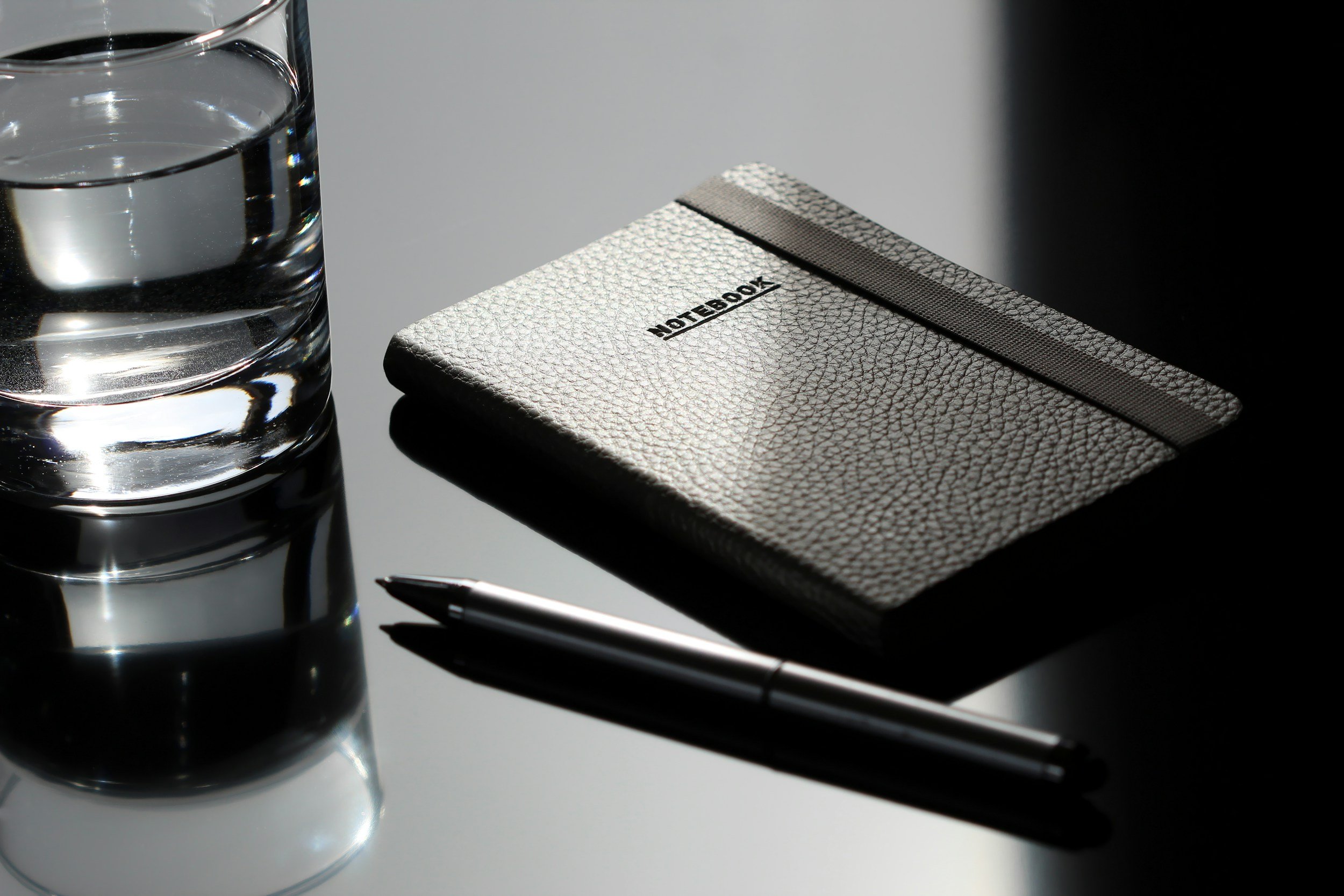 A glass of water, a silver notebook with a textured cover and black elastic band, a pen, and a silver pen are placed on a reflective surface.