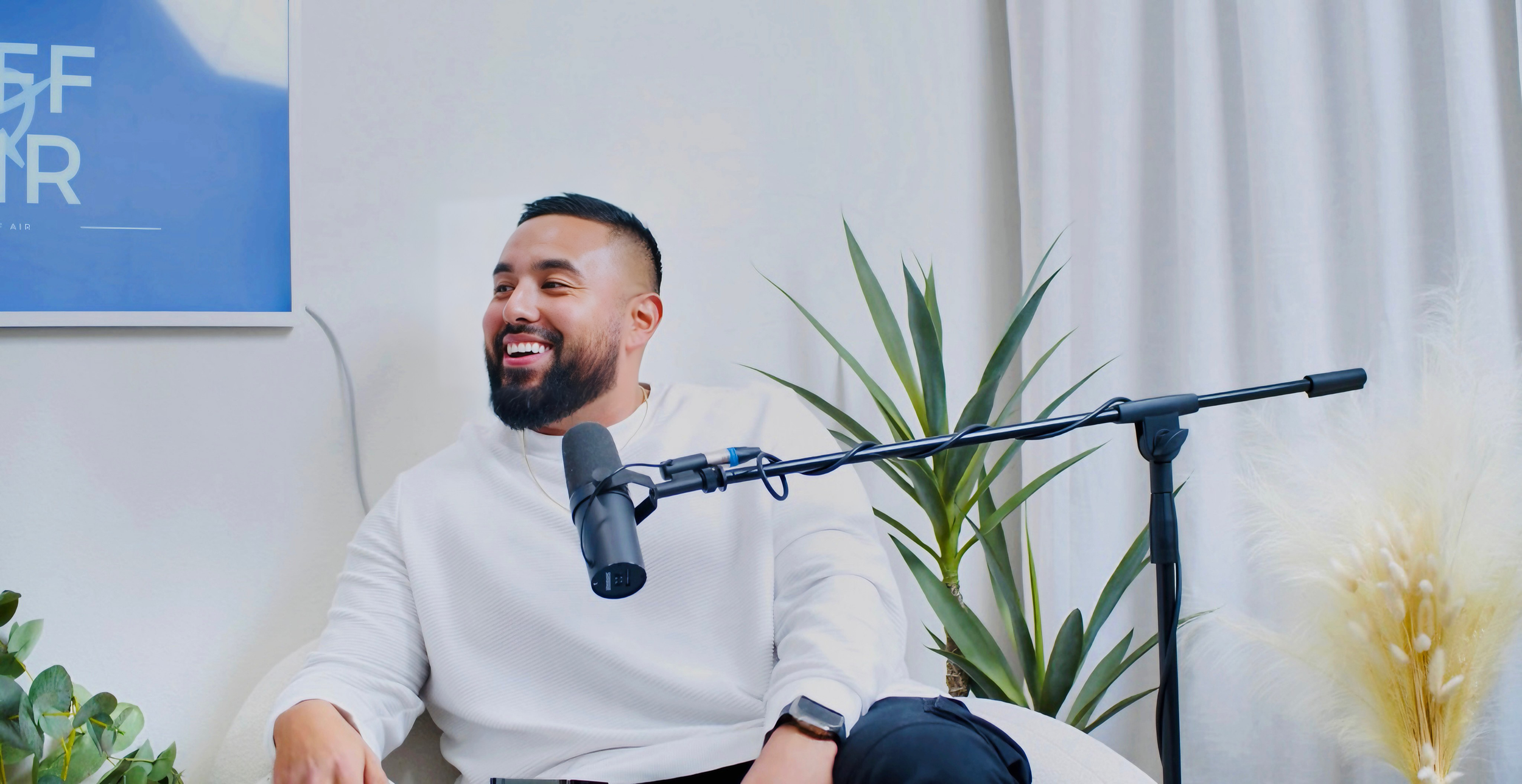 A man with a beard sitting on a white couch, smiling, with a microphone in front of him. There are plants and a blue sign on the wall behind him.
