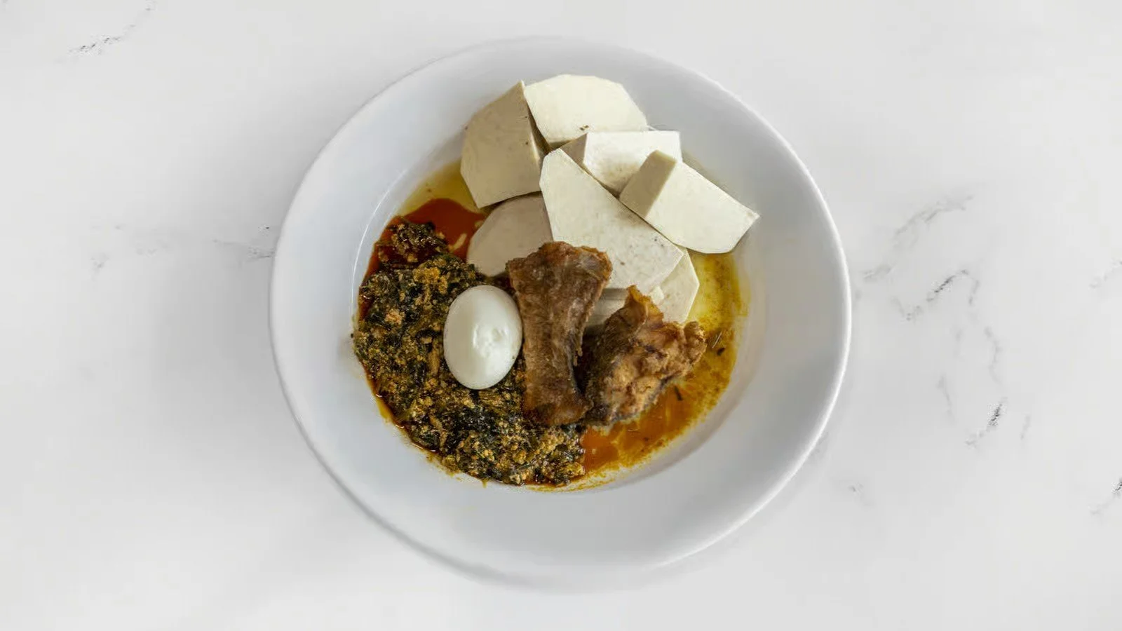 A white bowl containing traditional Nigerian dishes including boiled yam slices, stewed spinach, cooked goat meat, and a hard-boiled egg, with some palm oil or sauce.