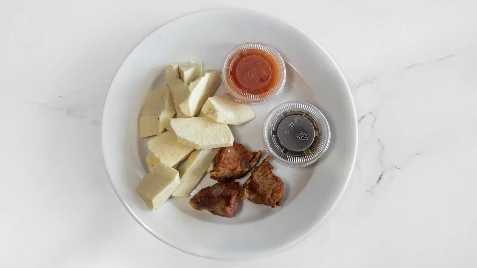 A white plate with chunks of boiled cassava, two pieces of cooked bacon, and two small containers of hot sauce and soy sauce on a white marble surface.