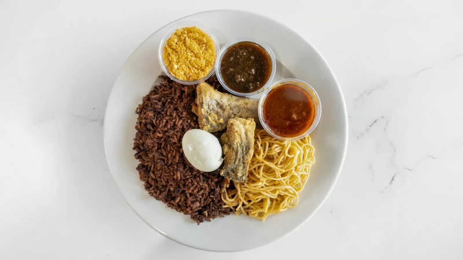 Plate of Nasi Lemak with rice, boiled egg, fried fish, noodles, and three small bowls of condiments and sauces.