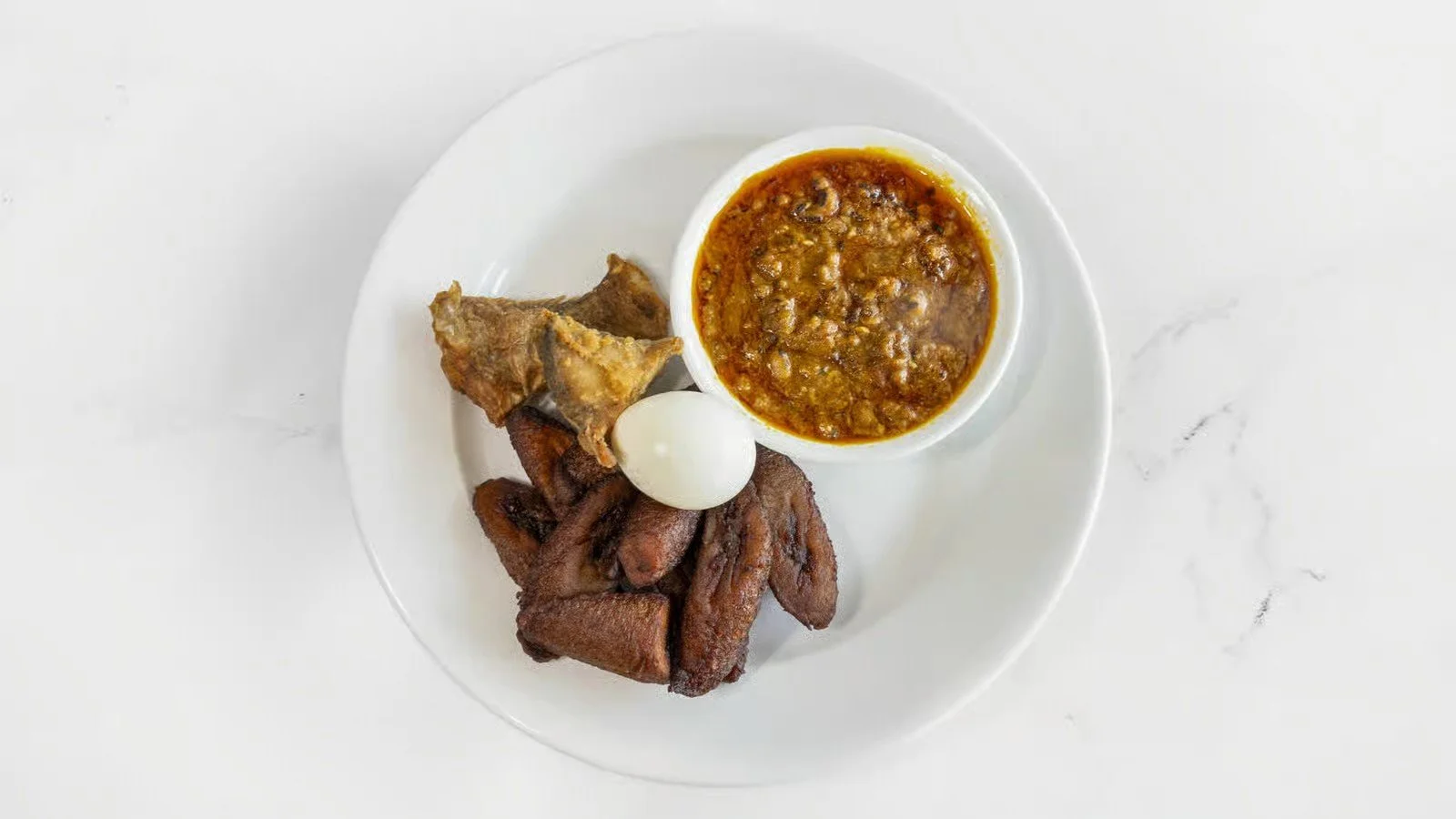 Plate of fried plantains, beef stew in a white bowl, and a boiled egg on a white surface.