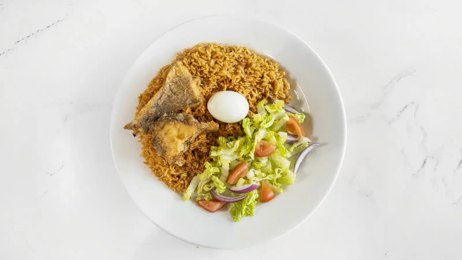 Plate of jollof rice with fried fish, boiled egg, and a side salad of lettuce, tomatoes, and onions.