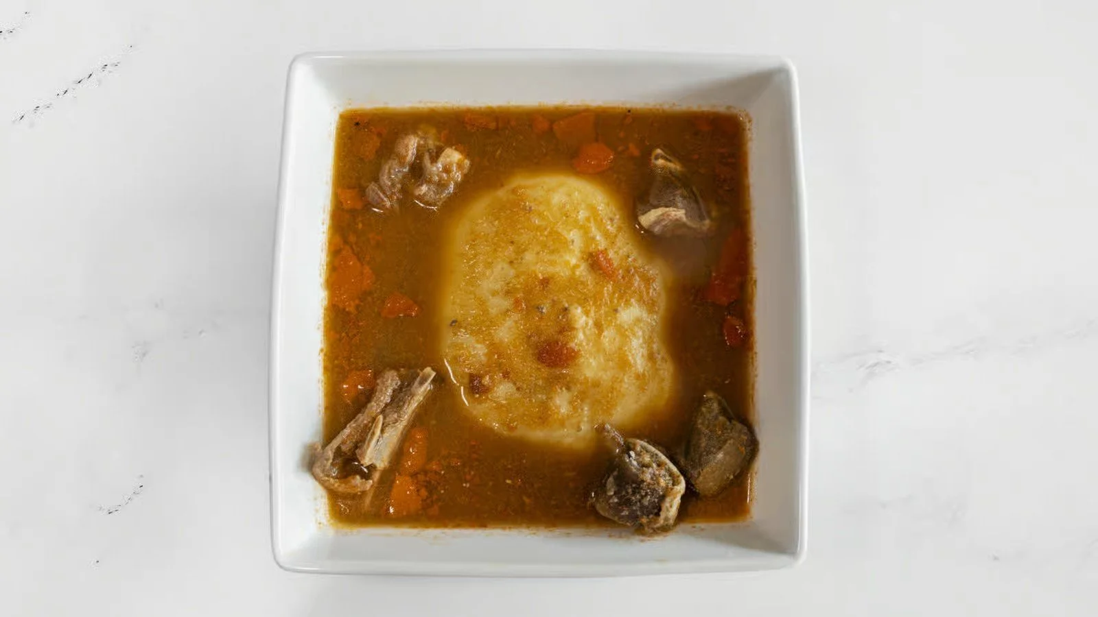 A bowl of beef stew with a dollop of mashed potatoes on top, garnished with chunks of beef and vegetables, in a white bowl on a white surface.