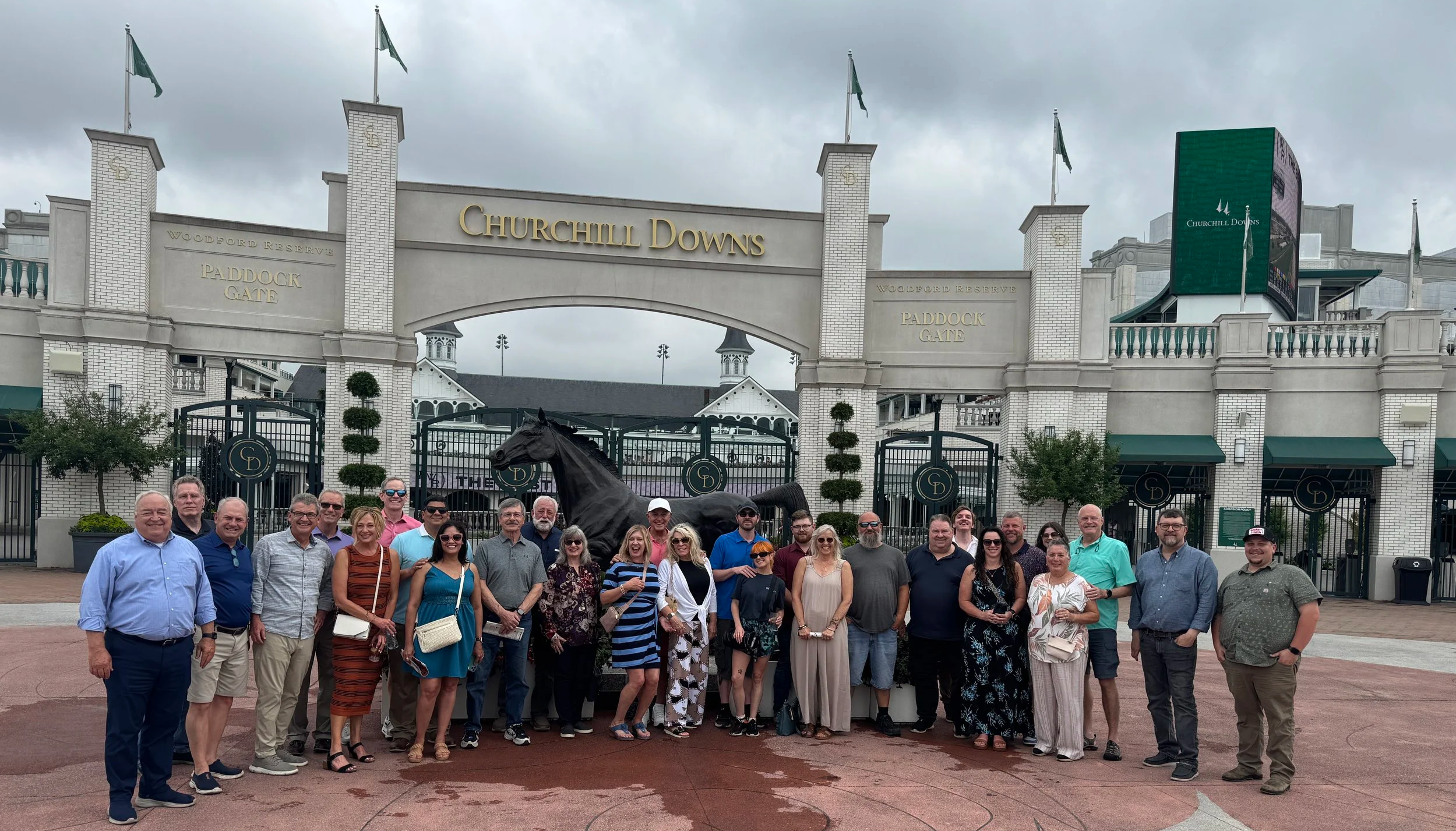A group of people posing in front of Churchill Downs racetrack entrance with the sign, a black horse statue, and a digital screen, on a cloudy day.