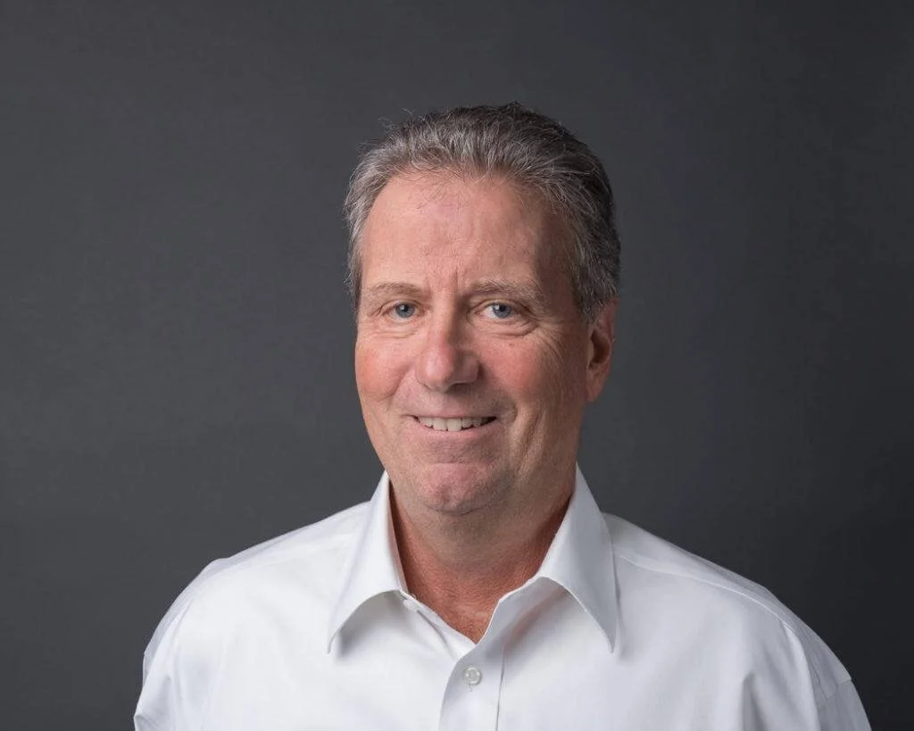 Rick Hirsh wearing a white collared shirt, smiling against a dark gray background.