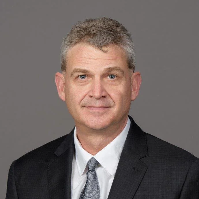 Portrait of a middle-aged man with short gray hair, wearing a black suit, white shirt, and patterned gray tie, against a plain gray background.
