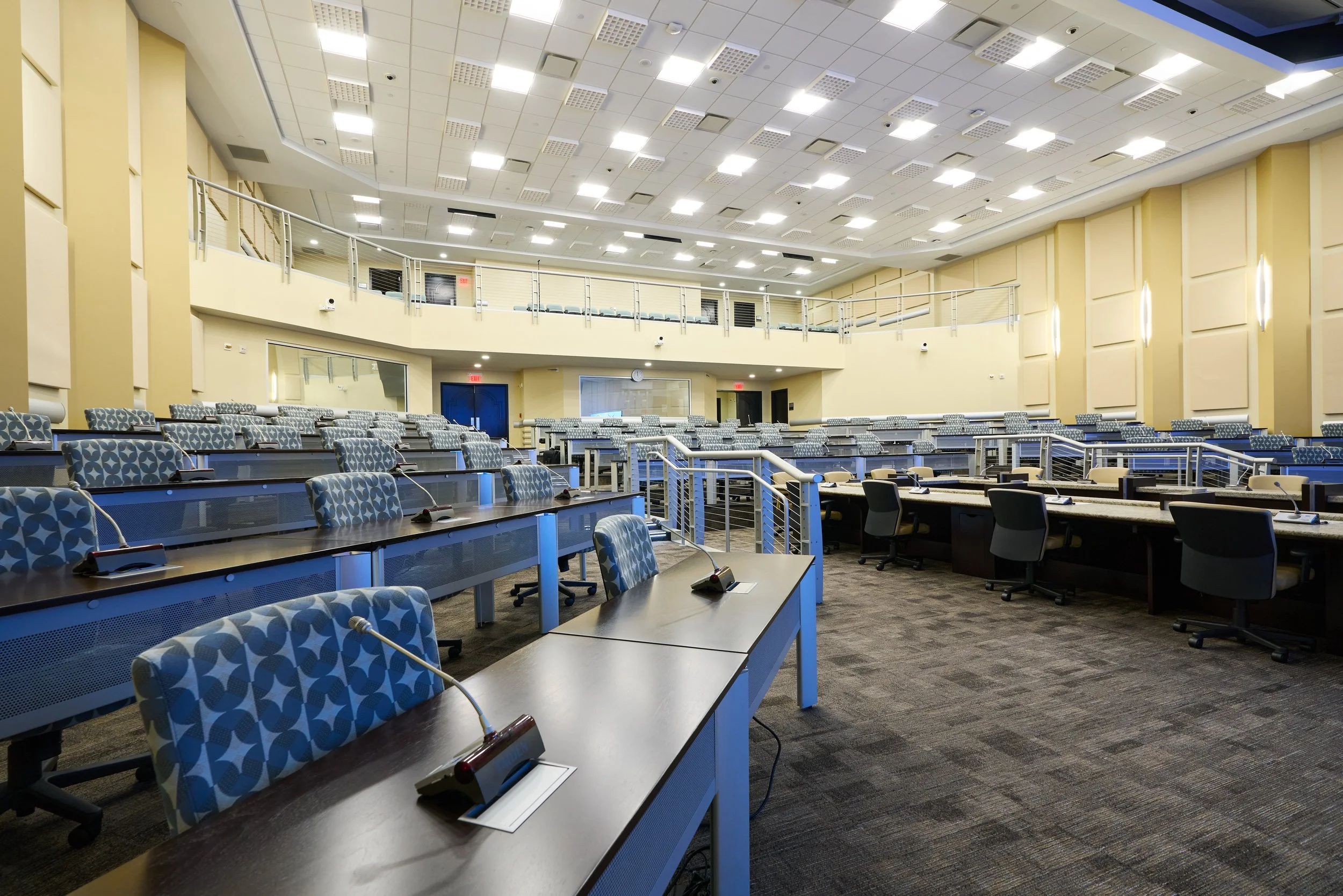 A large empty conference or lecture hall with multiple rows of tables and chairs, microphones on each table, and a balcony with additional seating. The hall has a high ceiling with numerous bright lights, beige walls with decorative paneling, and a carpeted floor.