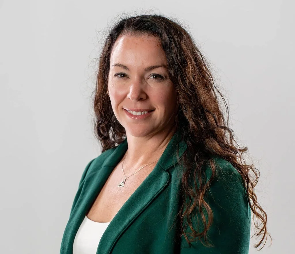 Julie Kolb, wearing a green blazer and white top, smiling against a plain light gray background.