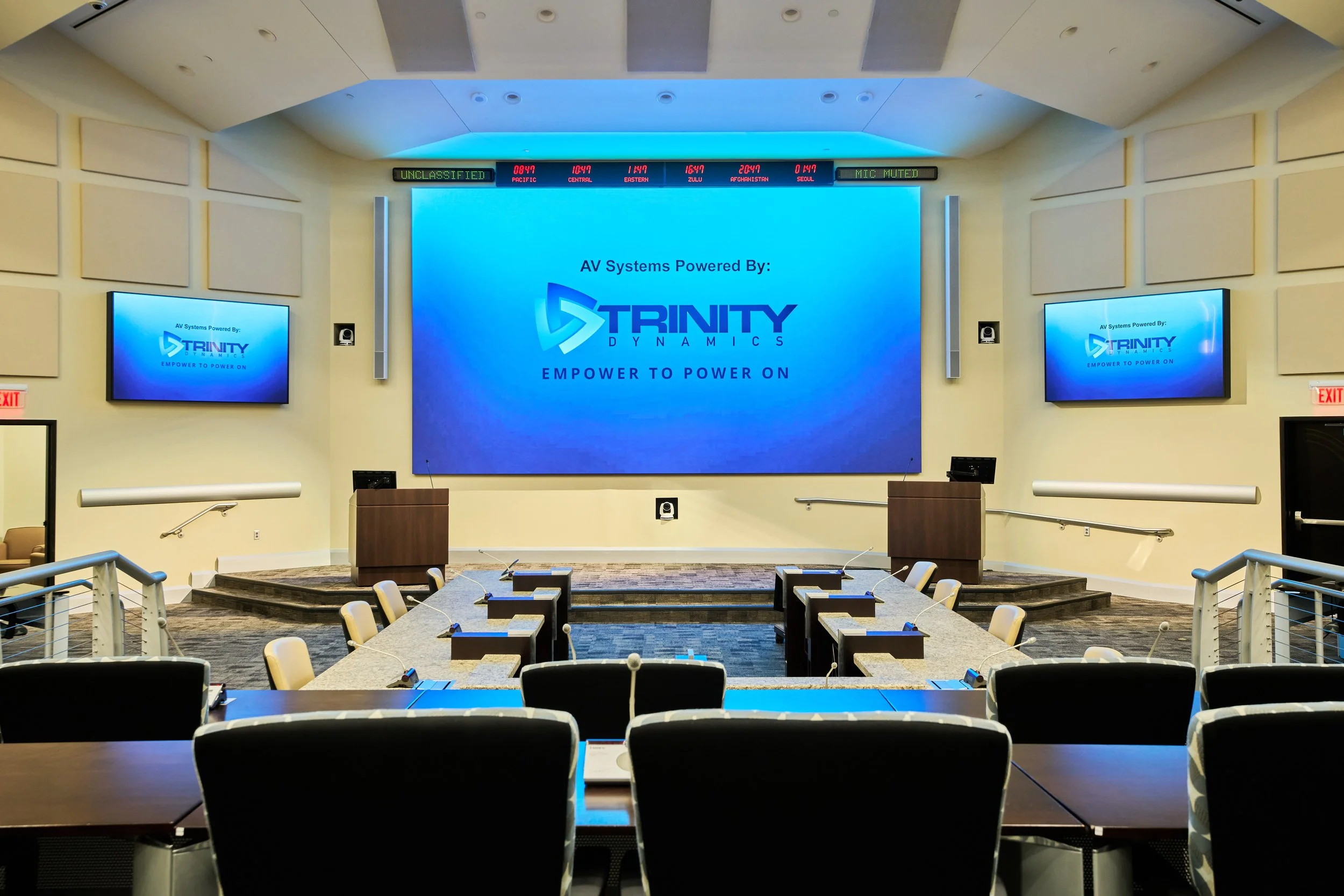Empty conference room with a stage, multiple monitors displaying Trinity Dynamics branding, and empty chairs and tables arranged for a meeting or presentation.