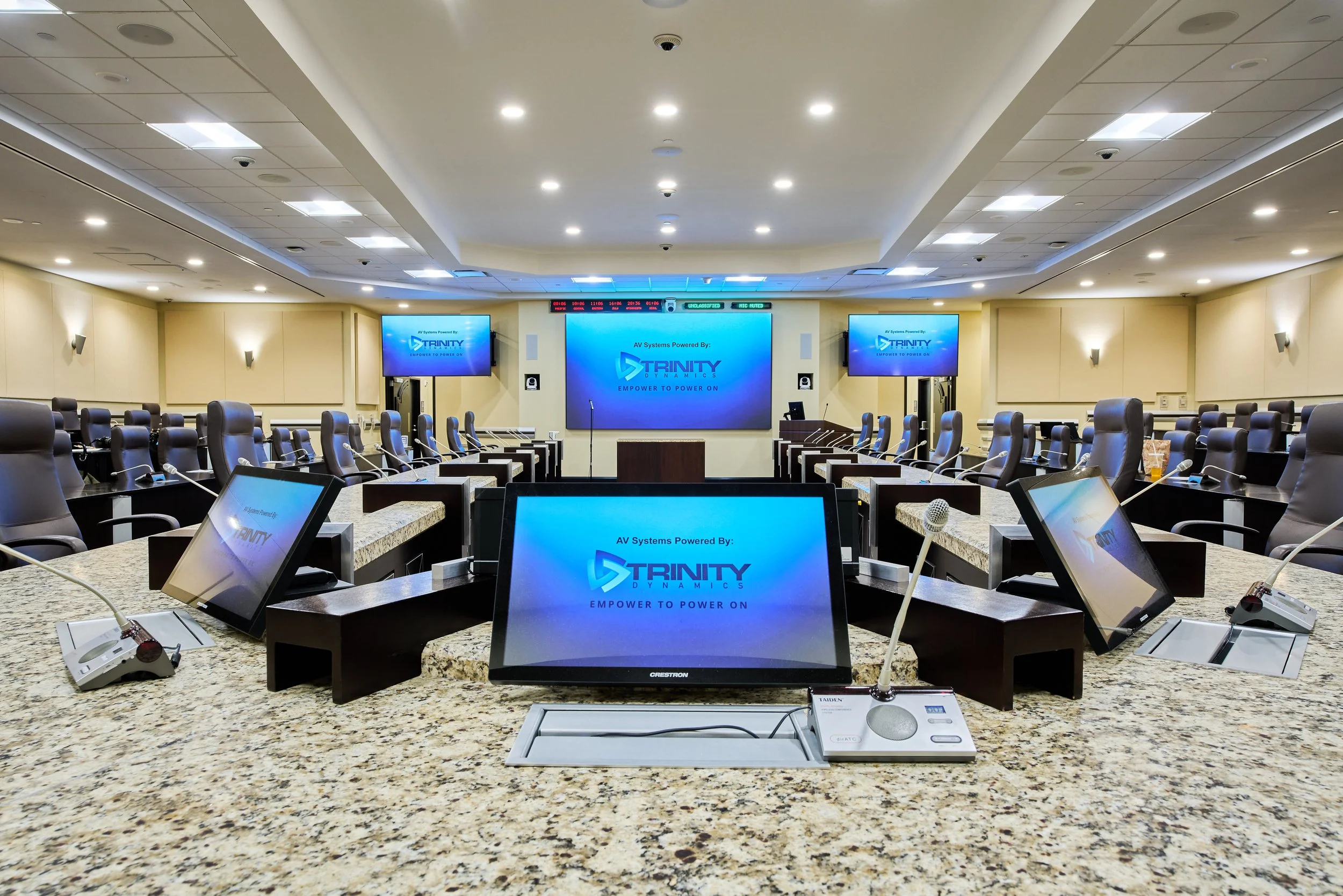 Empty conference room with large digital screens displaying 'Trinity Dynamics' logo, rows of black leather chairs, and microphones on the granite countertop with conference materials.
