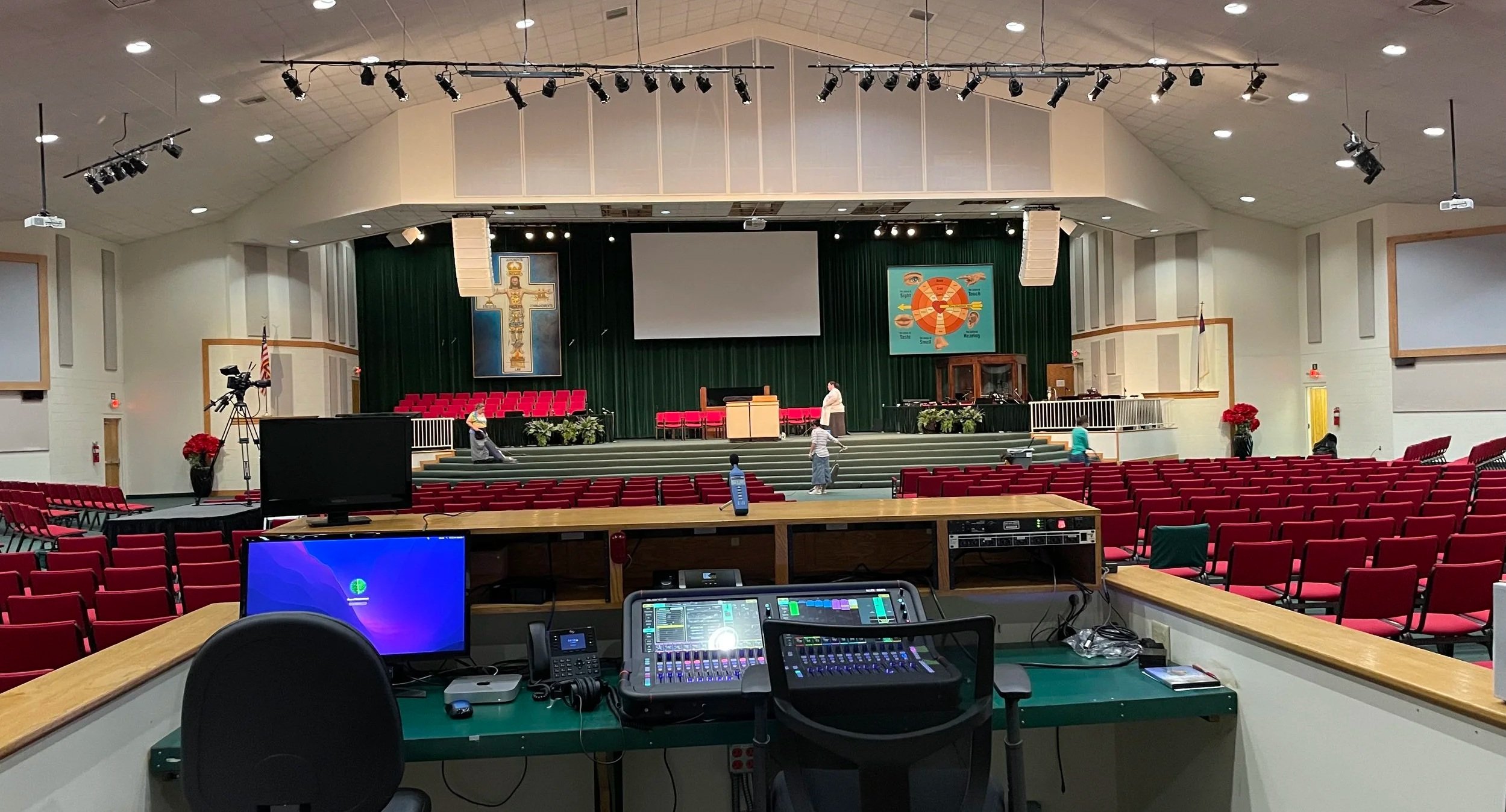 An auditorium or church stage with red chairs, green curtains, religious banners, and a podium, with people preparing for an event. The image is viewed from the control booth with audio and video equipment.