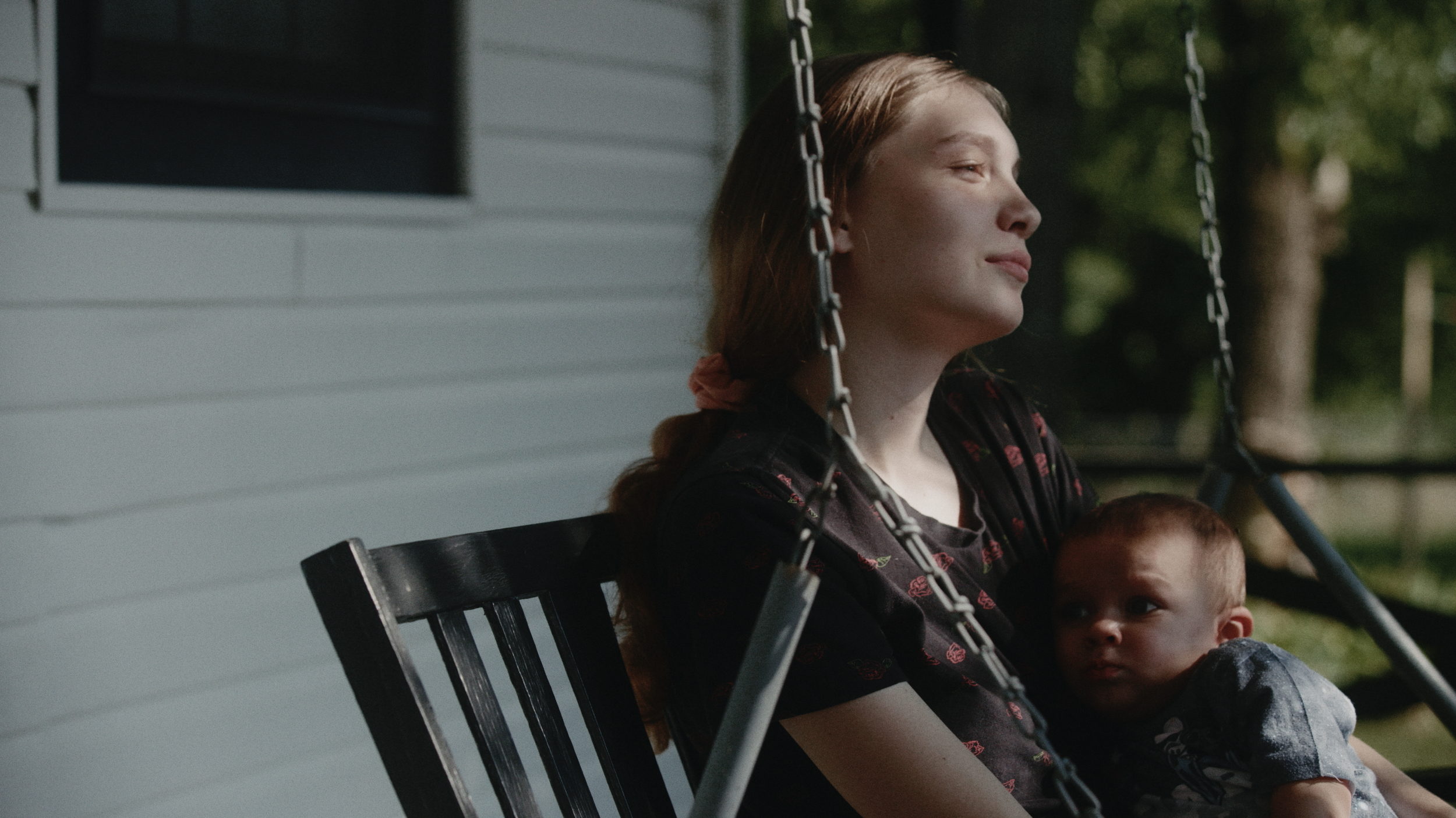 A woman with red hair and a young child sitting on a black porch swing with chains, outside a house with light blue siding, surrounded by trees.