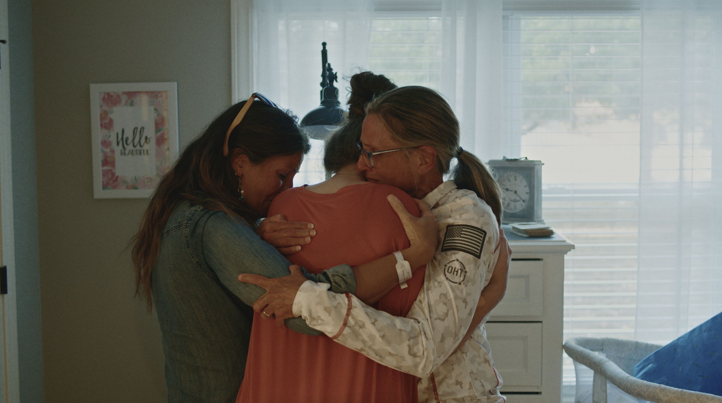 Three women, one in a U.S. Army camouflage uniform, embracing each other in a hospital room, showing support and comfort.