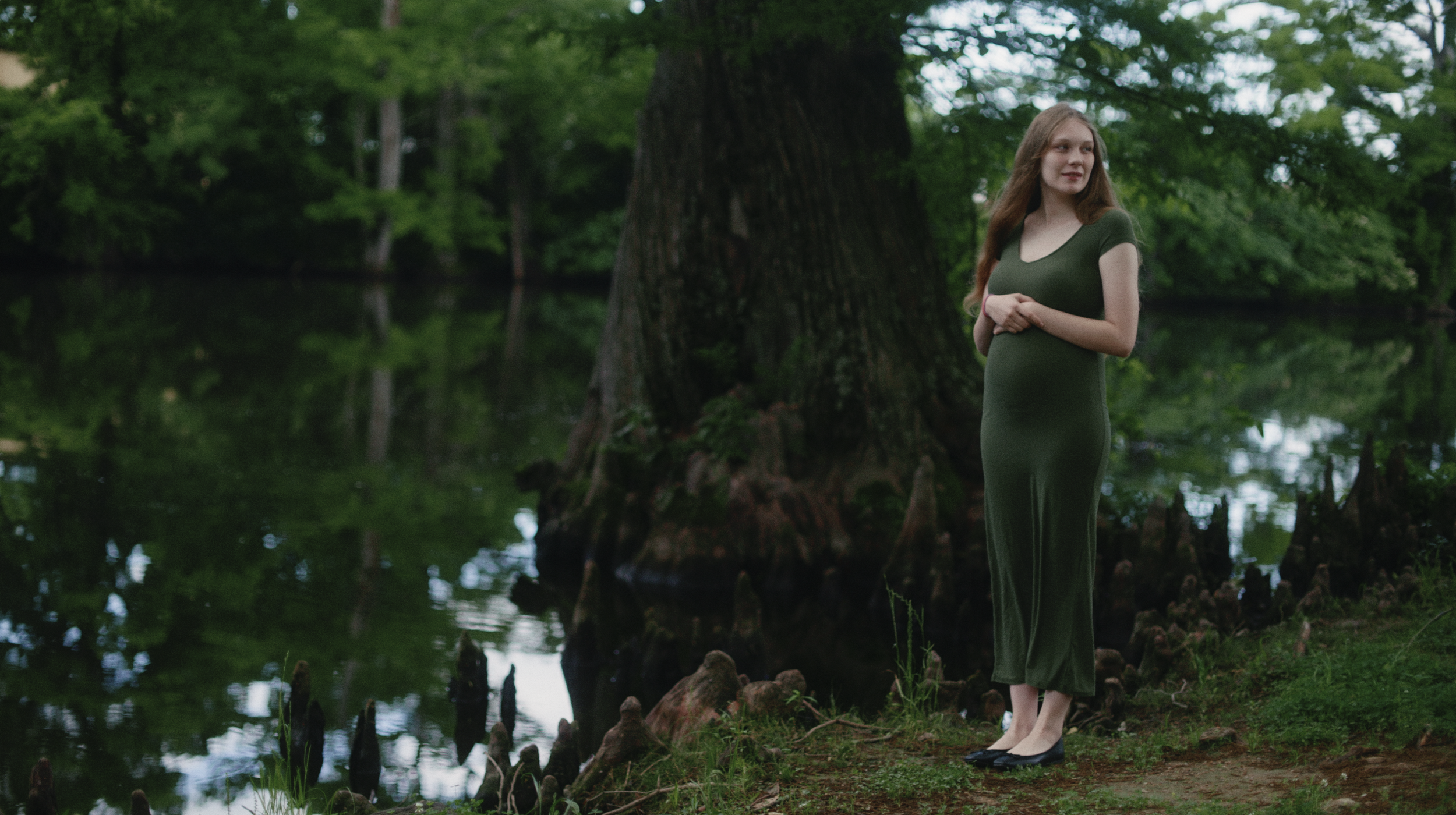 A woman in a long green dress standing by a large tree next to a body of water in a lush, green forest.