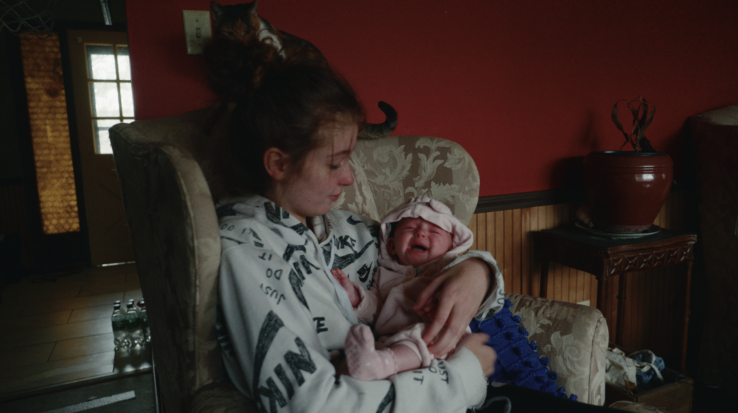 A young woman with closed eyes sitting on a floral patterned armchair, holding a crying newborn baby wrapped in a blanket, with a cat perched on her head.