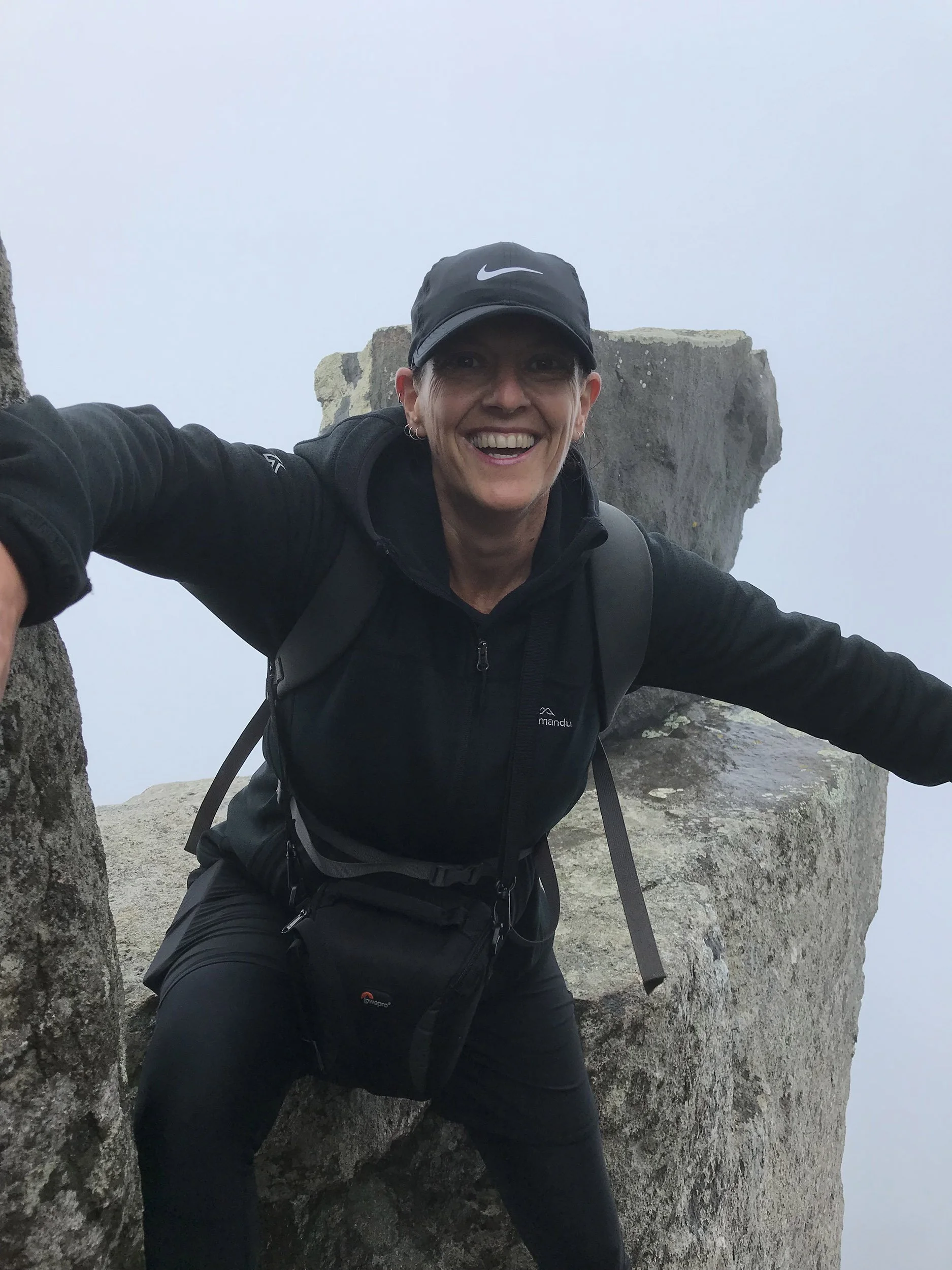 Person standing on a narrow rock ledge with arms outstretched between two large rock formations in foggy, dramatic windy mountain conditions