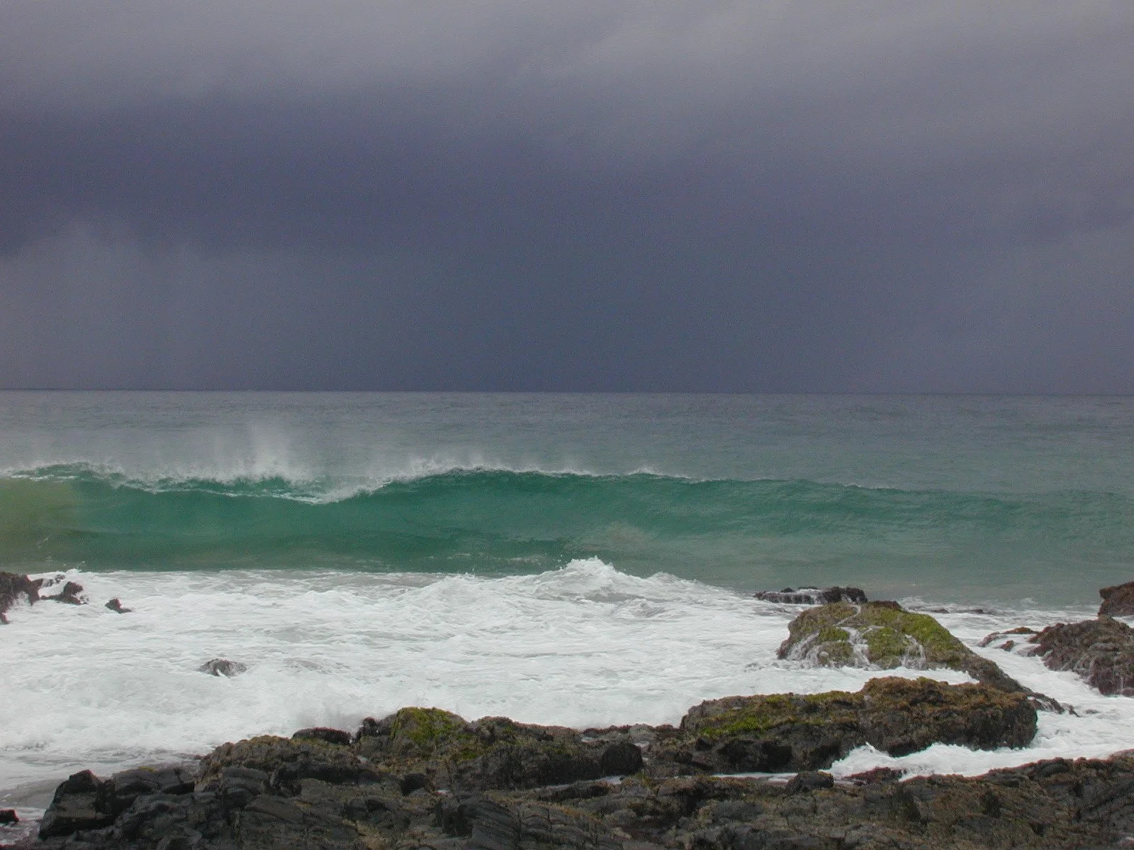 “Stormy coastal scene with dark clouds over turbulent green waves crashing against moss‑covered rocks on the shoreline