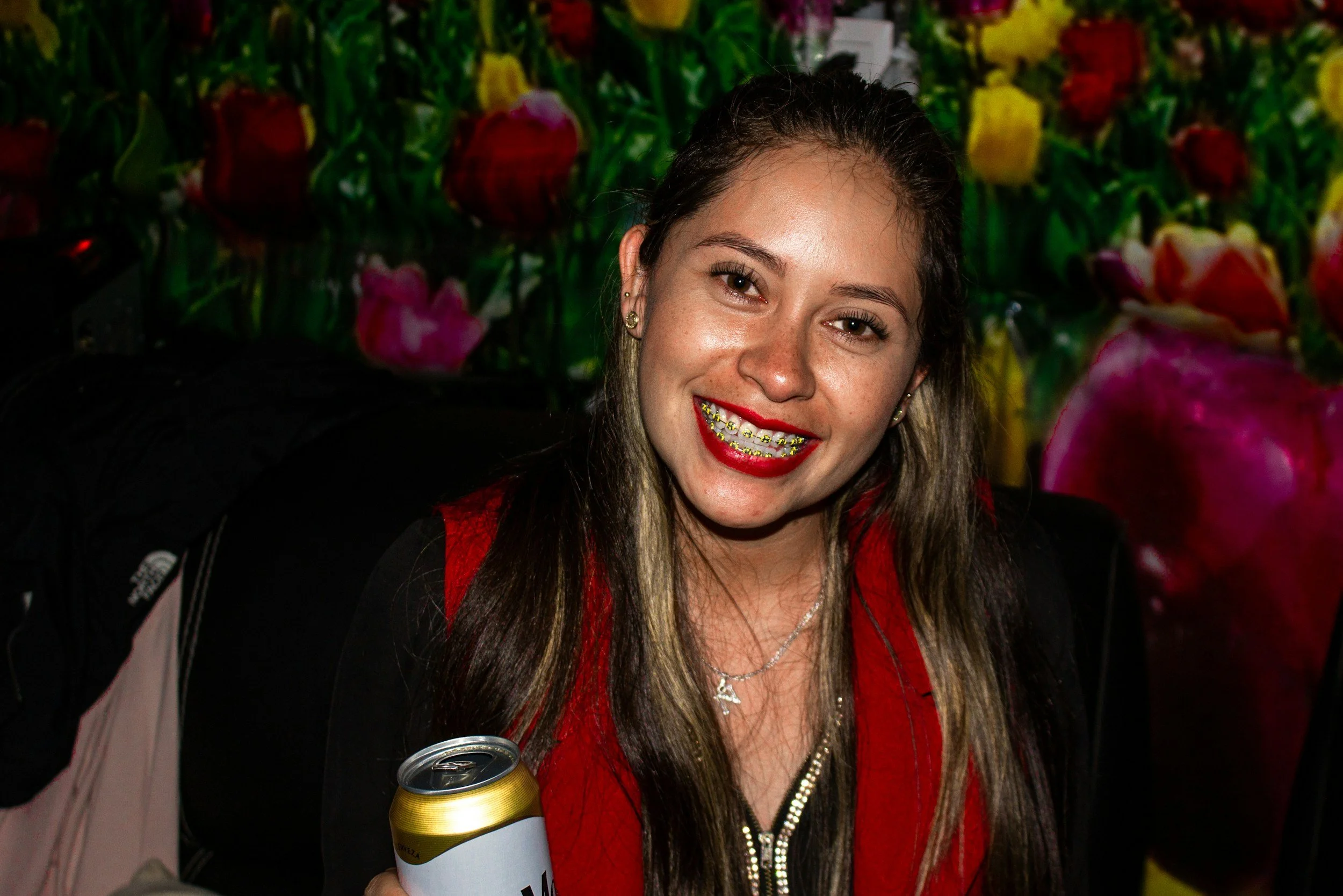 A young woman smiling with gold braces, holding a can of beer, with a colorful floral background.