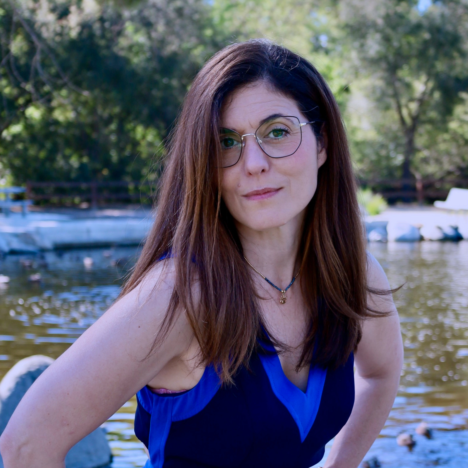 A woman with long brown hair and glasses posing outdoors near a body of water, with trees in the background.