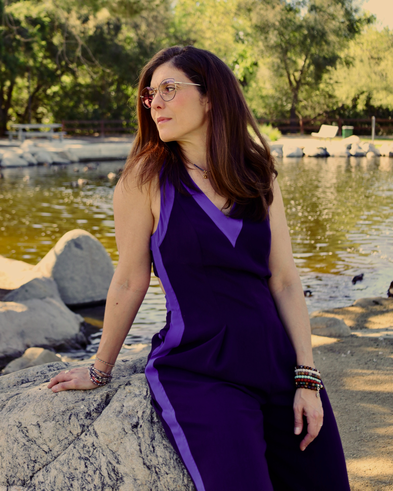 A woman with long, dark hair and glasses, wearing a purple sleeveless jumpsuit, sitting on a large rock by a pond with trees and benches in the background.