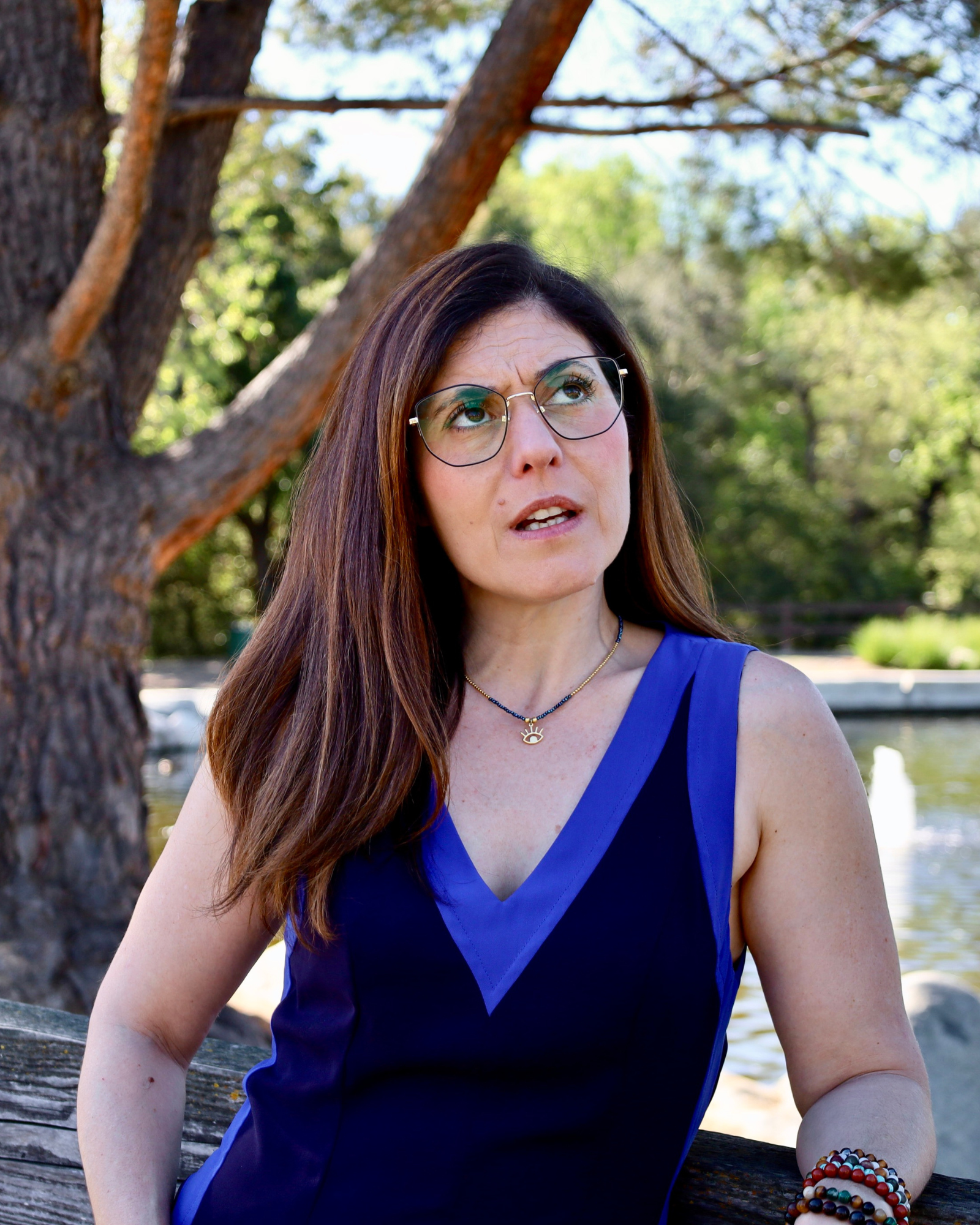 A woman with brown hair, glasses, and a blue sleeveless top standing outdoors near a tree and a body of water, with a confused or questioning expression on her face.