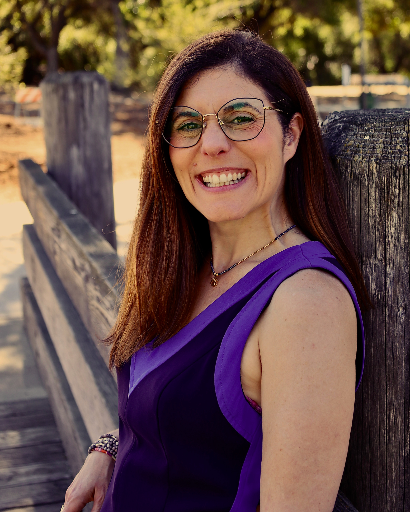 A woman with long brown hair wearing glasses and a purple sleeveless dress, smiling while sitting on a wooden bench outdoors on a sunny day.