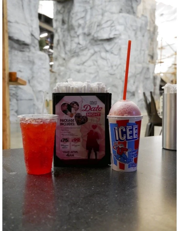 A red drink in a clear plastic cup, an icee with a red straw in a striped cup, and a promotional flyer for a date night package on a dark table, with a rocky background and seating area.