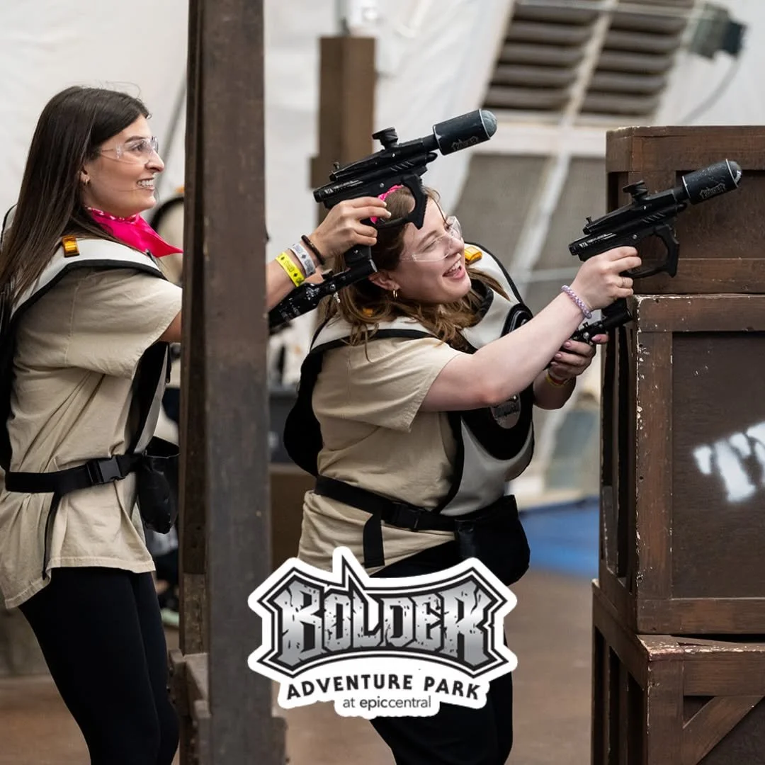 Two women engaged in laser tag at Bolder Adventure Park, with one woman crouching behind cover and aiming a laser gun, while the other stands behind her, both wearing safety goggles and beige vests.