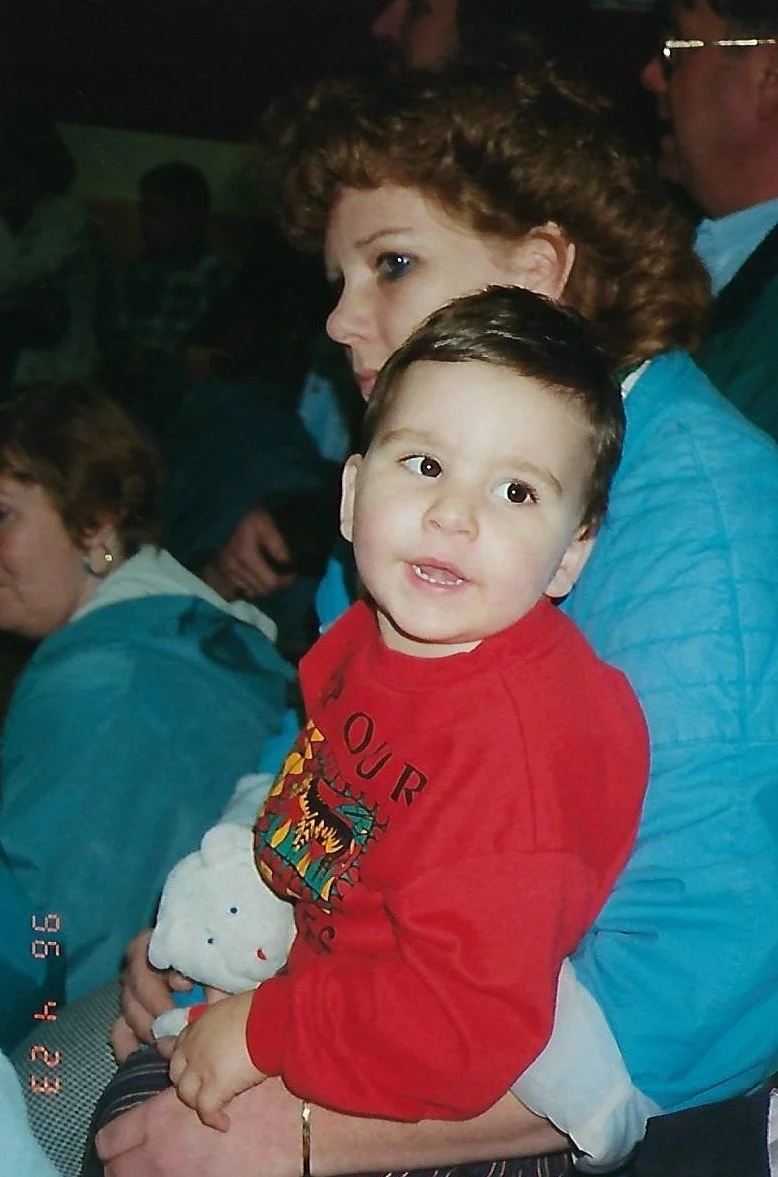 Young boy in a red shirt sitting on an adult's lap holding a white stuffed animal, with adults and other children in a dimly lit setting.