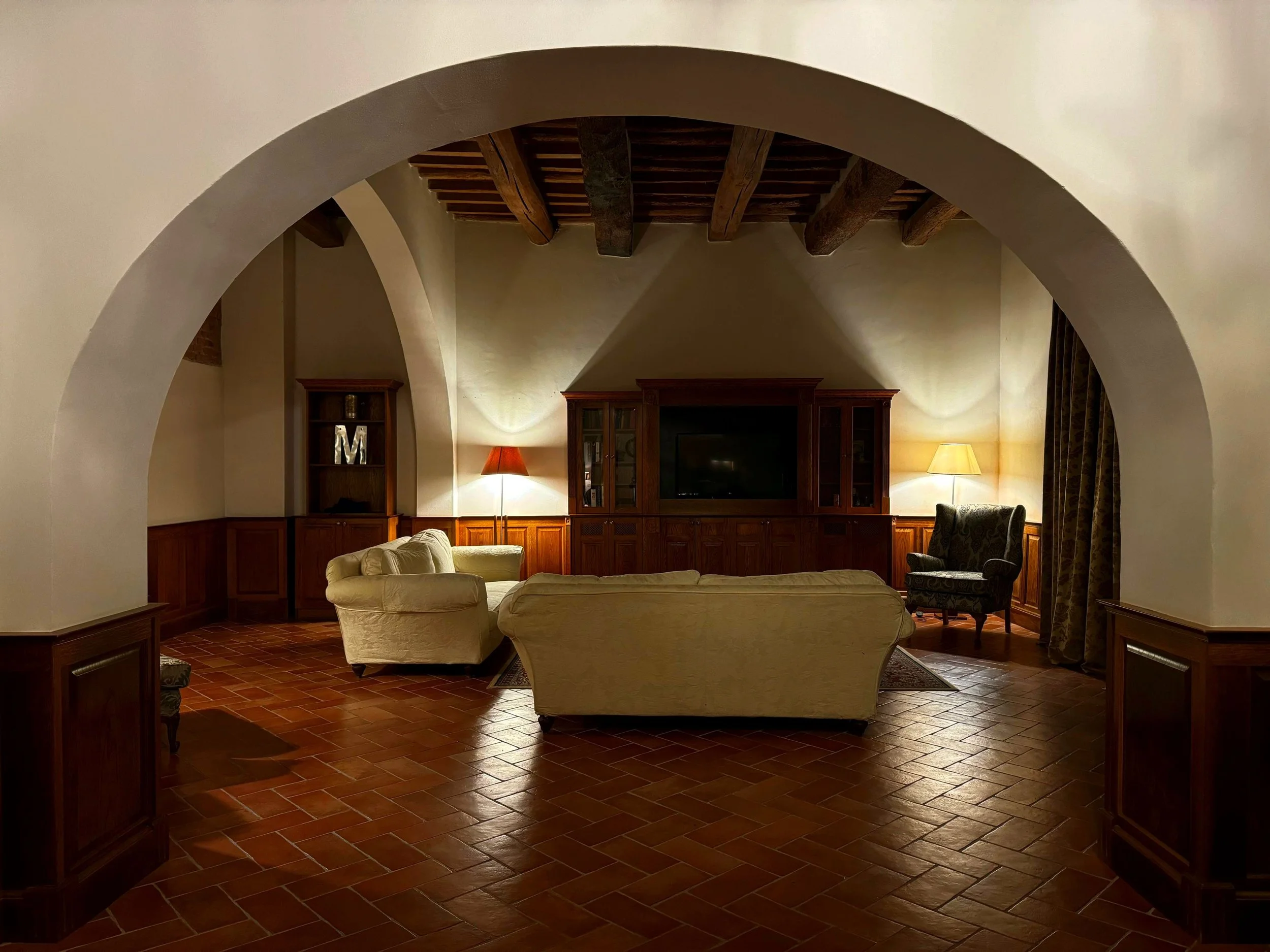 Living room with arched doorways, wooden ceiling beams, beige couches, a wooden entertainment center, and lamps.